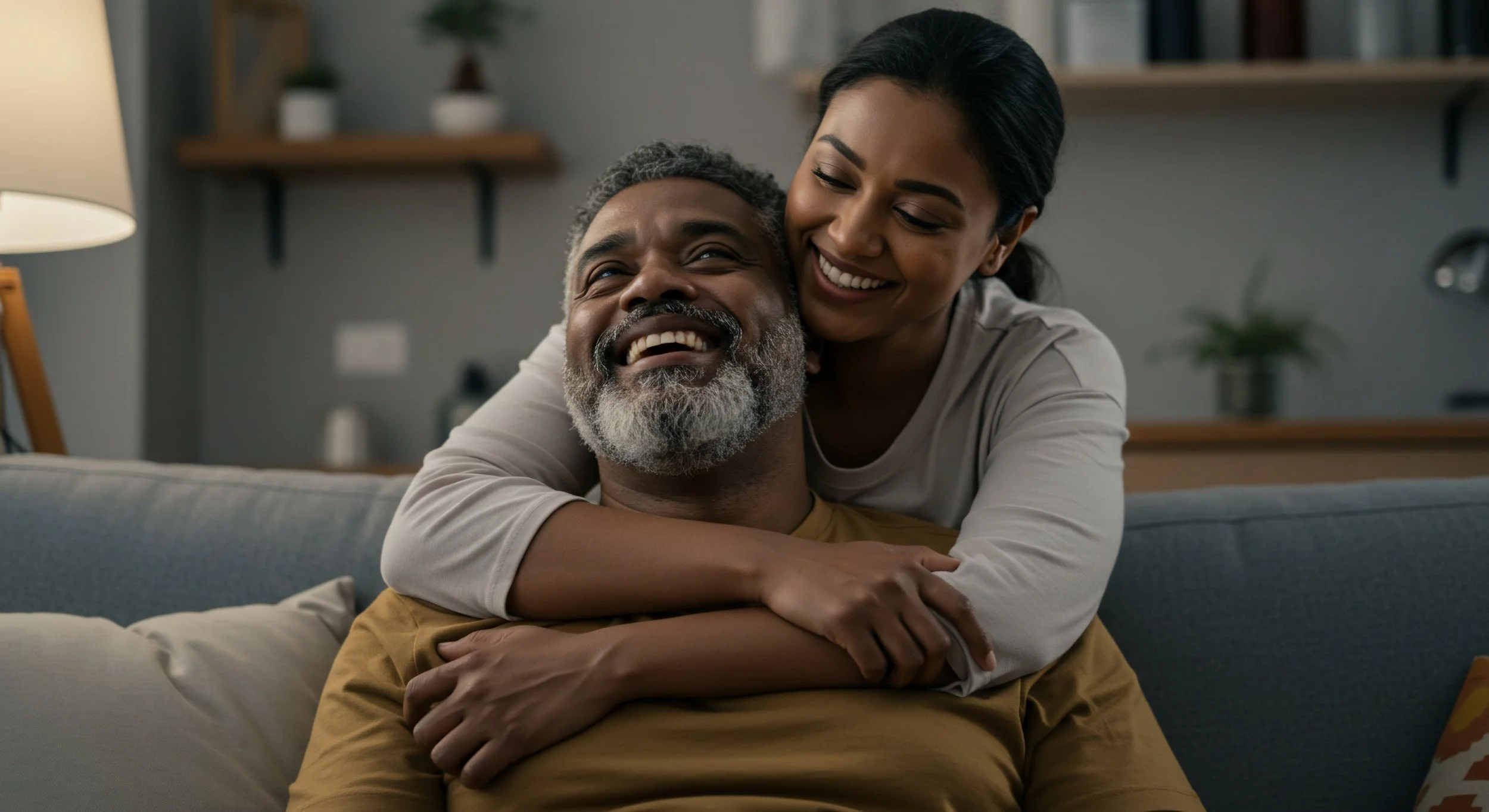 A woman hugging a man from behind on a couch, both smiling and laughing warmly.