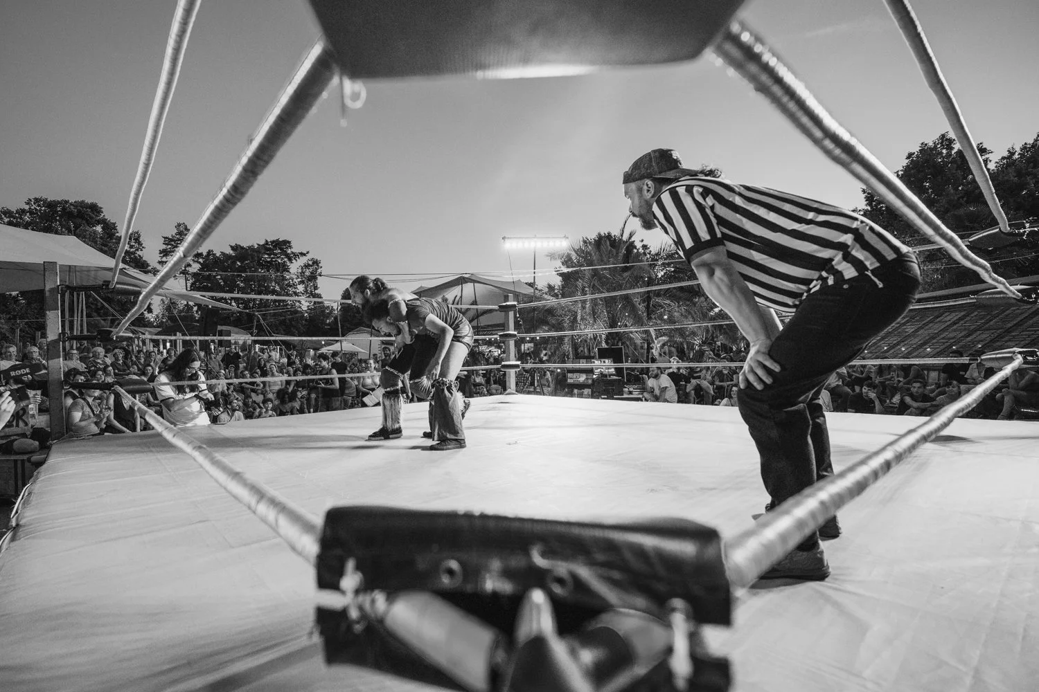 Black and white photo of an outdoor wrestling match viewed from the corner of the ring, with two female wrestlers bending over and a referee watching closely.
