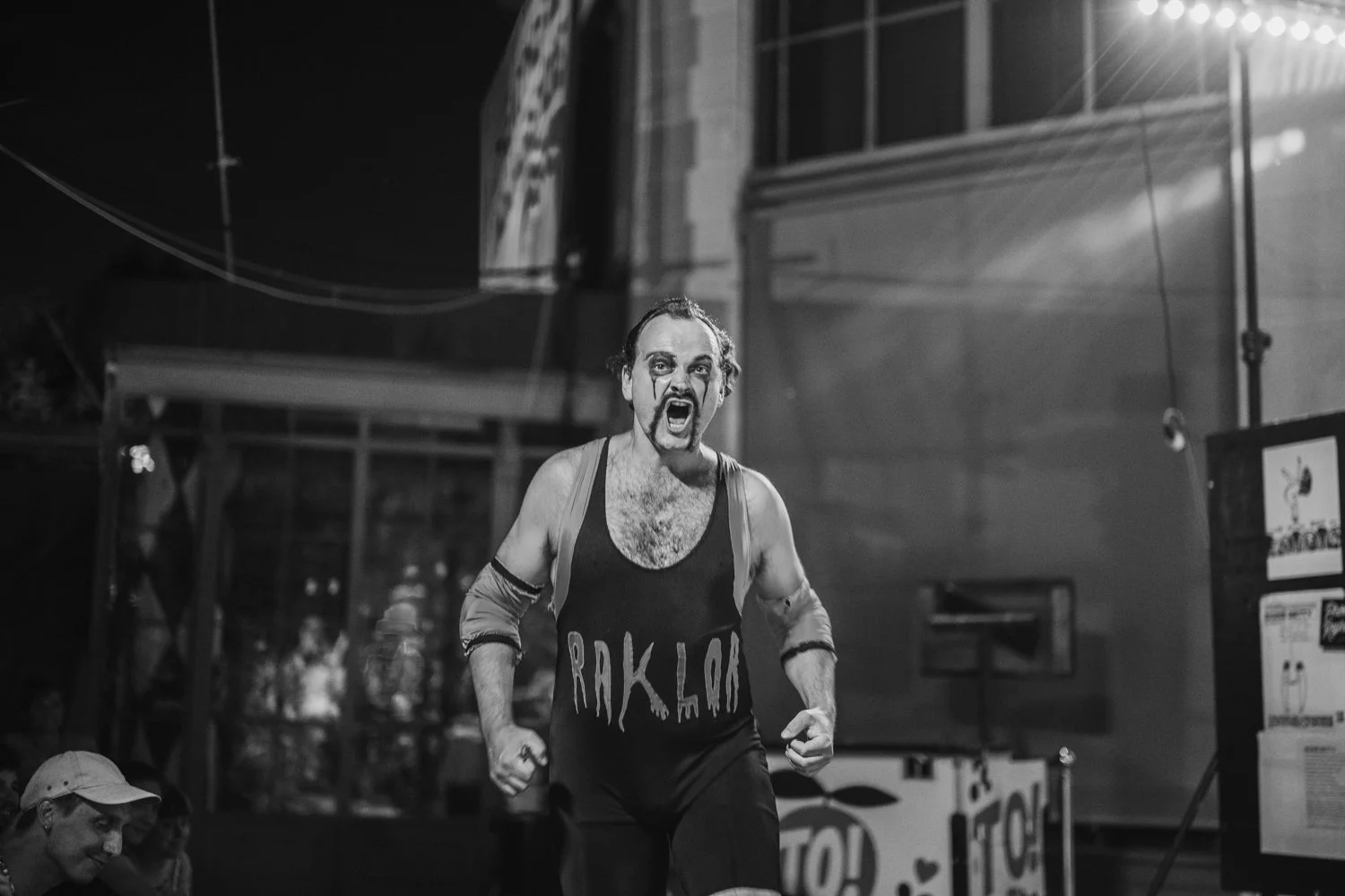 A man with makeup on his face, wearing a sleeveless shirt with the word 'RAKELAN' on it, appears to be shouting or yelling while running in an indoor area, possibly during a performance or event, with some people and signs visible in the background.