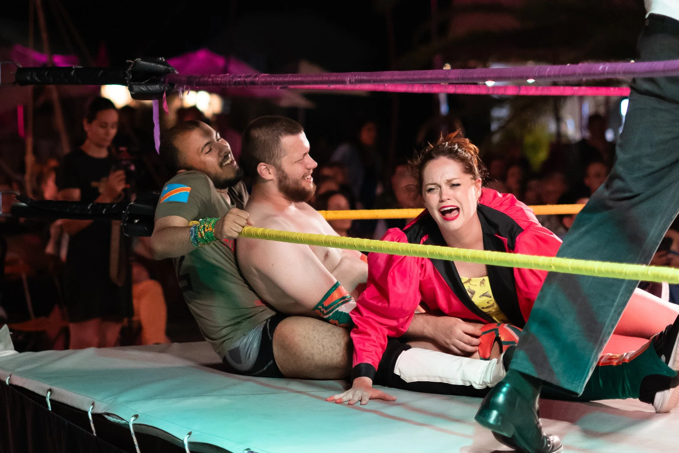 Two men and a woman engaged in a playful wrestling match inside a wrestling ring with yellow ropes, with a crowd watching in the background at night.