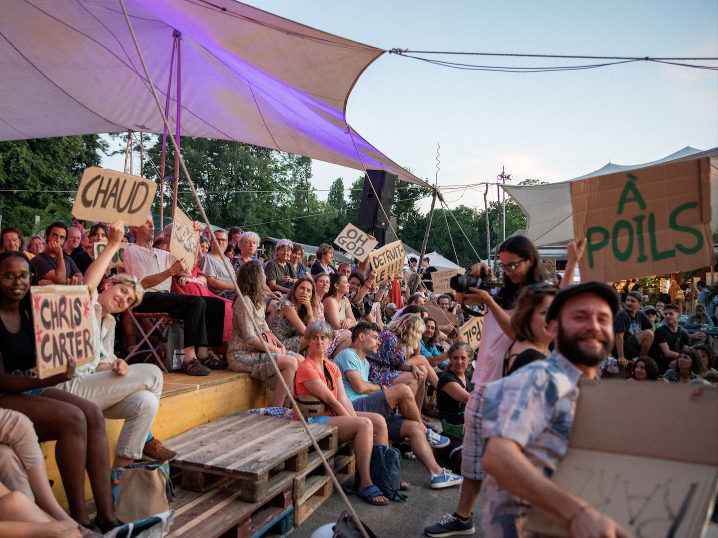 People gathered at an outdoor event, sitting on benches and holding handmade signs with names and messages, under large canopy tents during sunset.