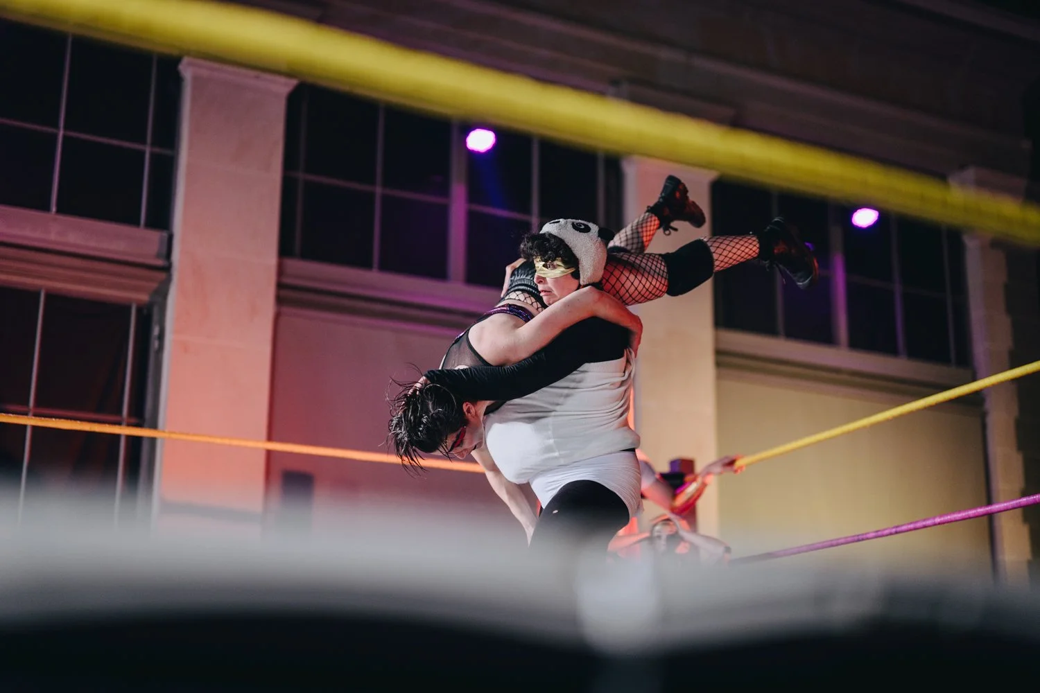 Two women wrestling in a ring with colorful ropes, one lifting the other in a move, inside a building with large windows and colorful lighting.