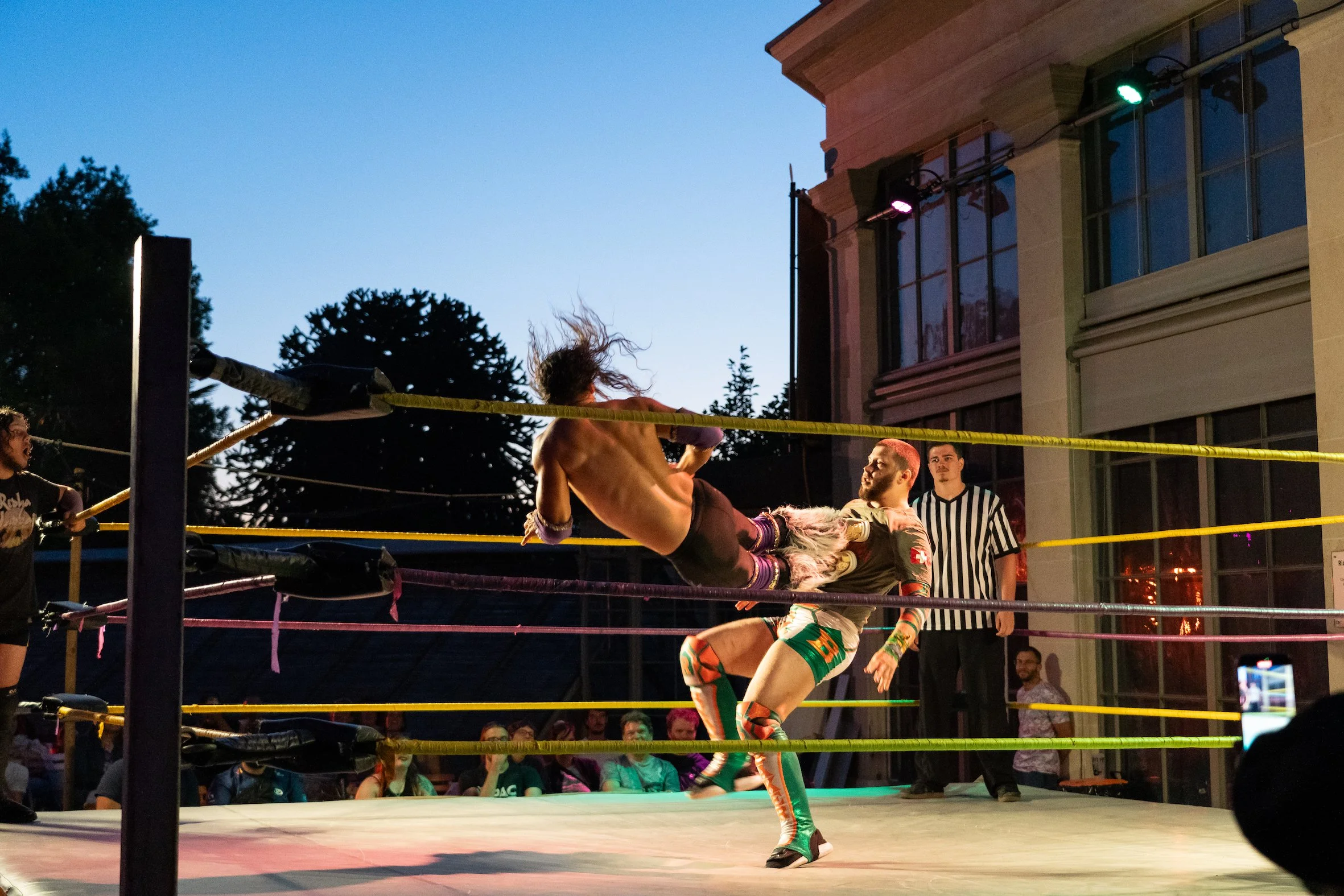 A professional wrestling match taking place outdoors during sunset. A wrestler is executing a kick to another wrestler, who is mid-air with long hair. Spectators and a referee are watching the action.