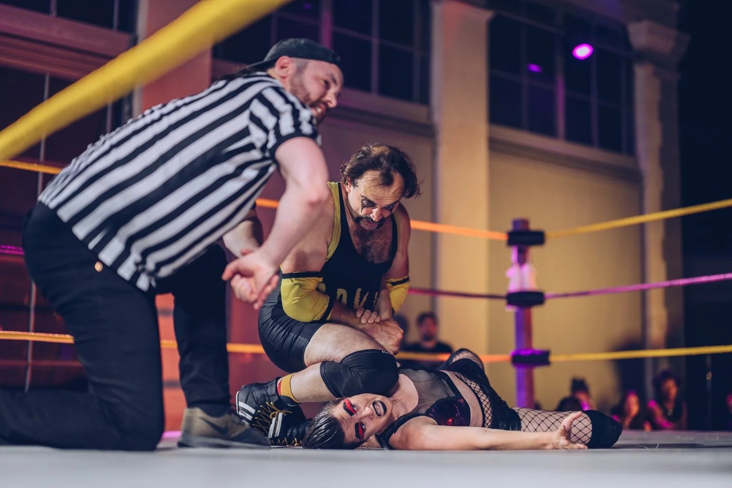 A wrestling match with a male wrestler pinning a female opponent on the mat with the referee nearby observing the match.