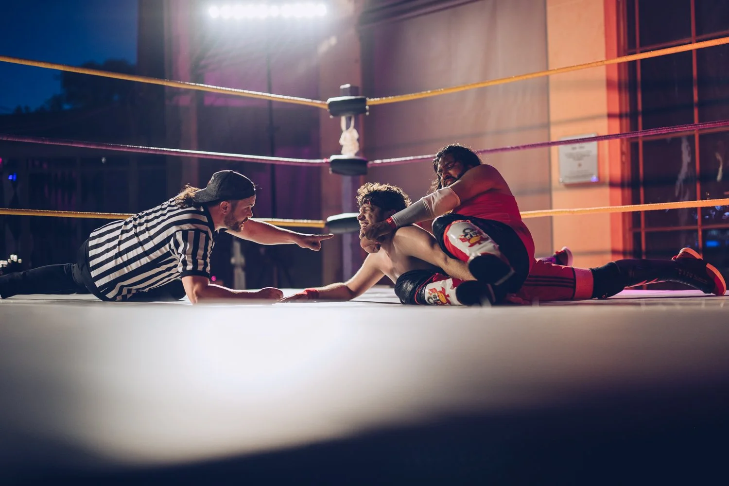 Two wrestlers on the mat in a wrestling ring, one pinning the other, with a referee reaching towards them, in an indoor wrestling arena.