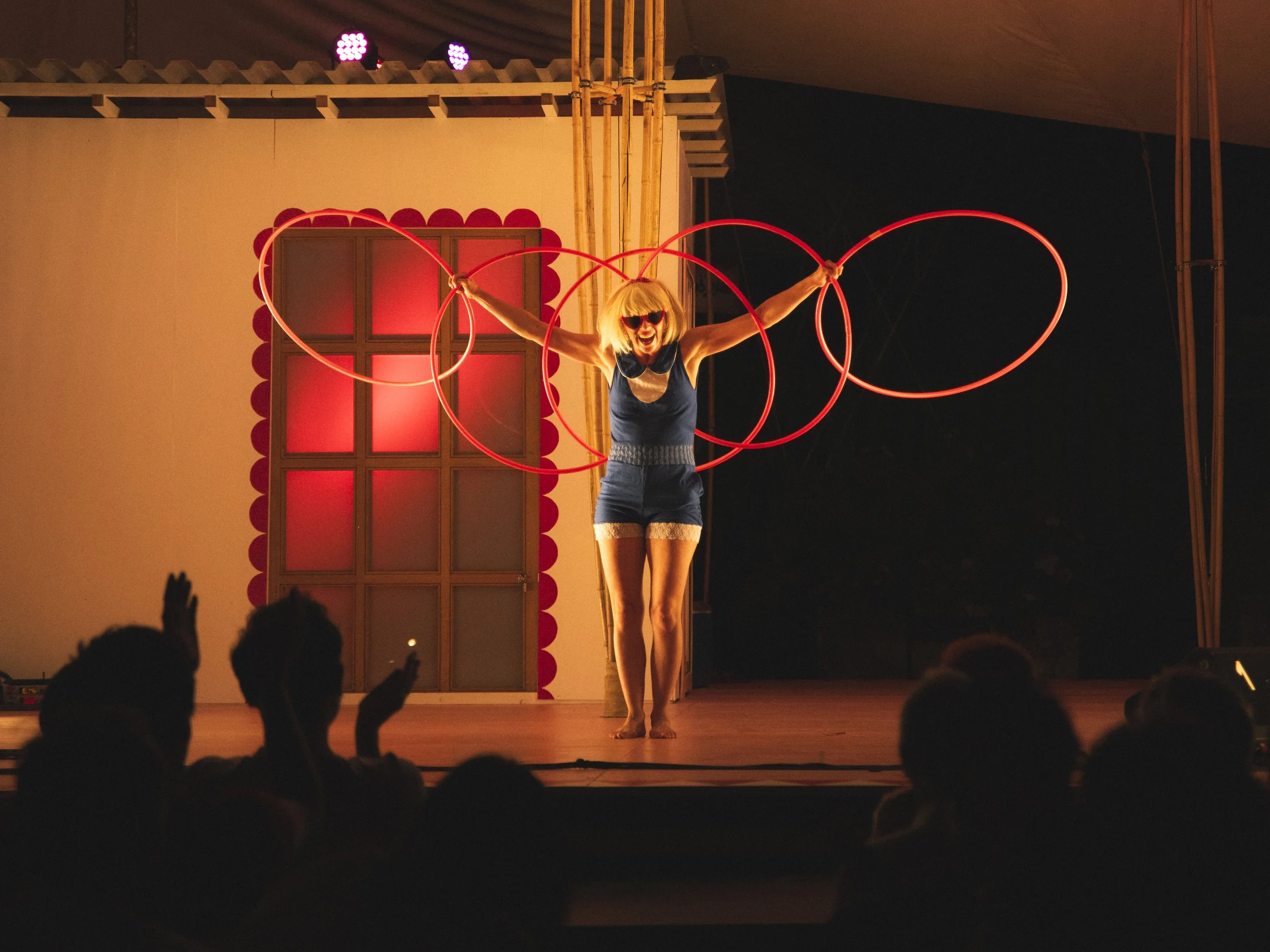 Performer with blonde hair and sunglasses spinning red hoops on stage, audience clapping and cheering