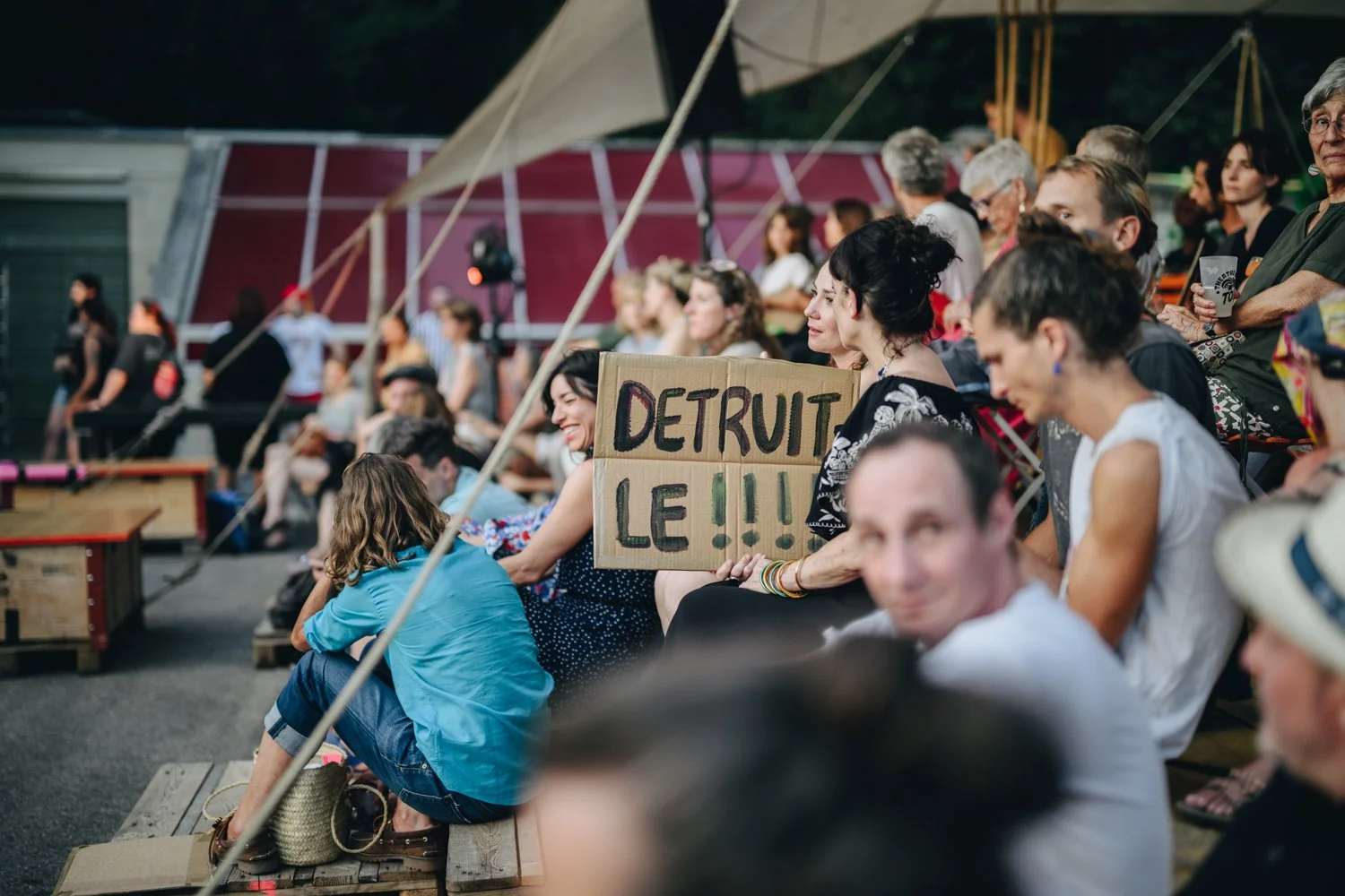 Crowd of people sitting in an outdoor theater, one woman holding a cardboard sign that reads 'DETRUIT LE!!'