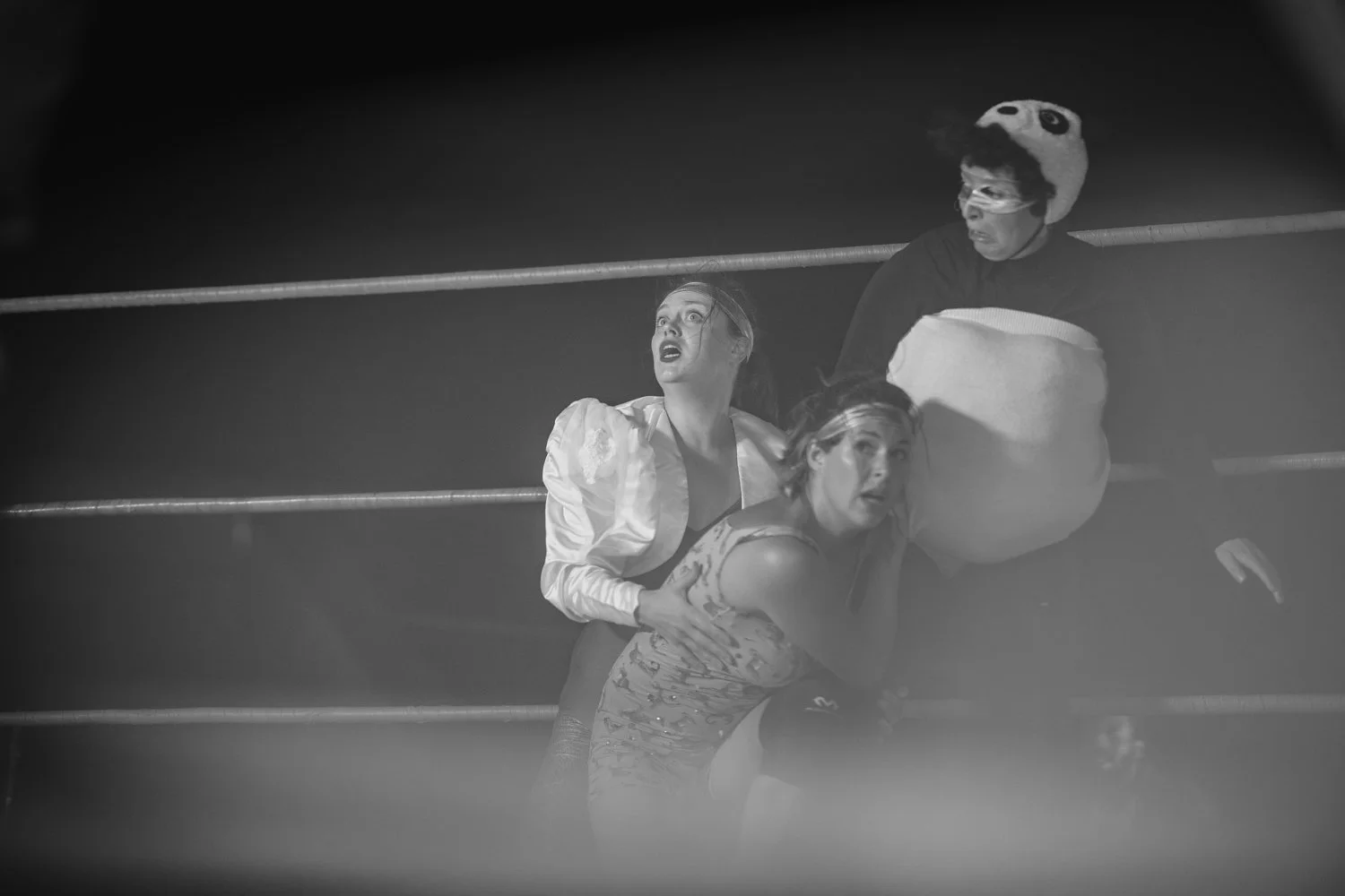 Three performers in costumes on a stage, with two women in vintage-style dresses and a person in a panda costume, appearing surprised or frightened.