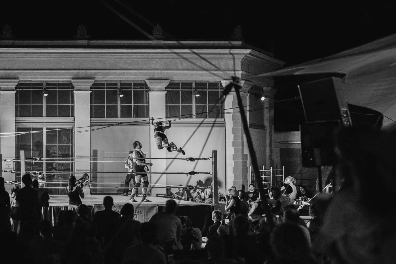 Black and white photo of a wrestling match with a wrestler flying through the air, in front of an audience during a night event, outdoors, in front of a large building with tall windows.