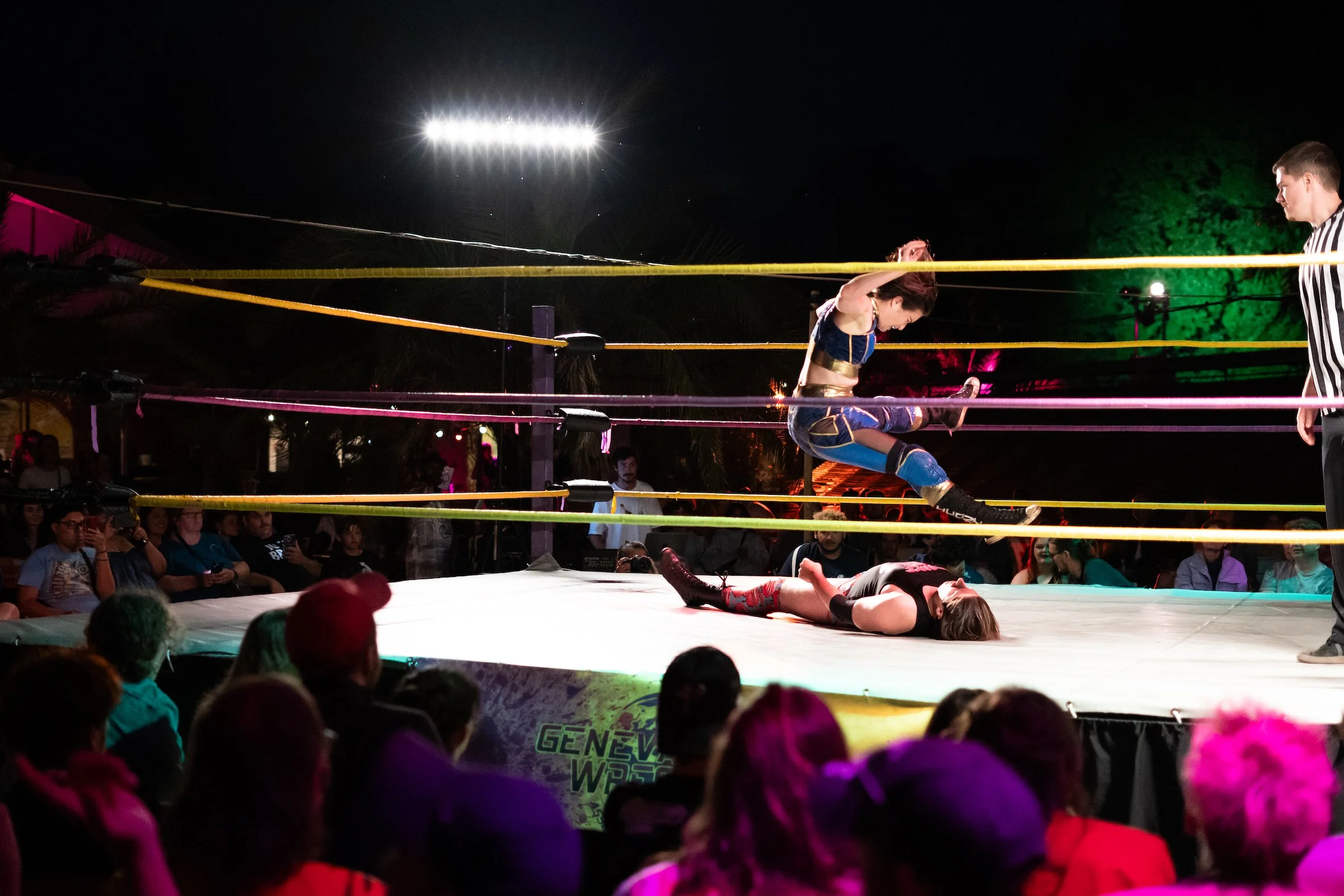 Professional women's wrestling match with a woman mid-air executing a move on her opponent lying on the mat, crowd watching, referee standing nearby, colorful lighting, outdoor night event.