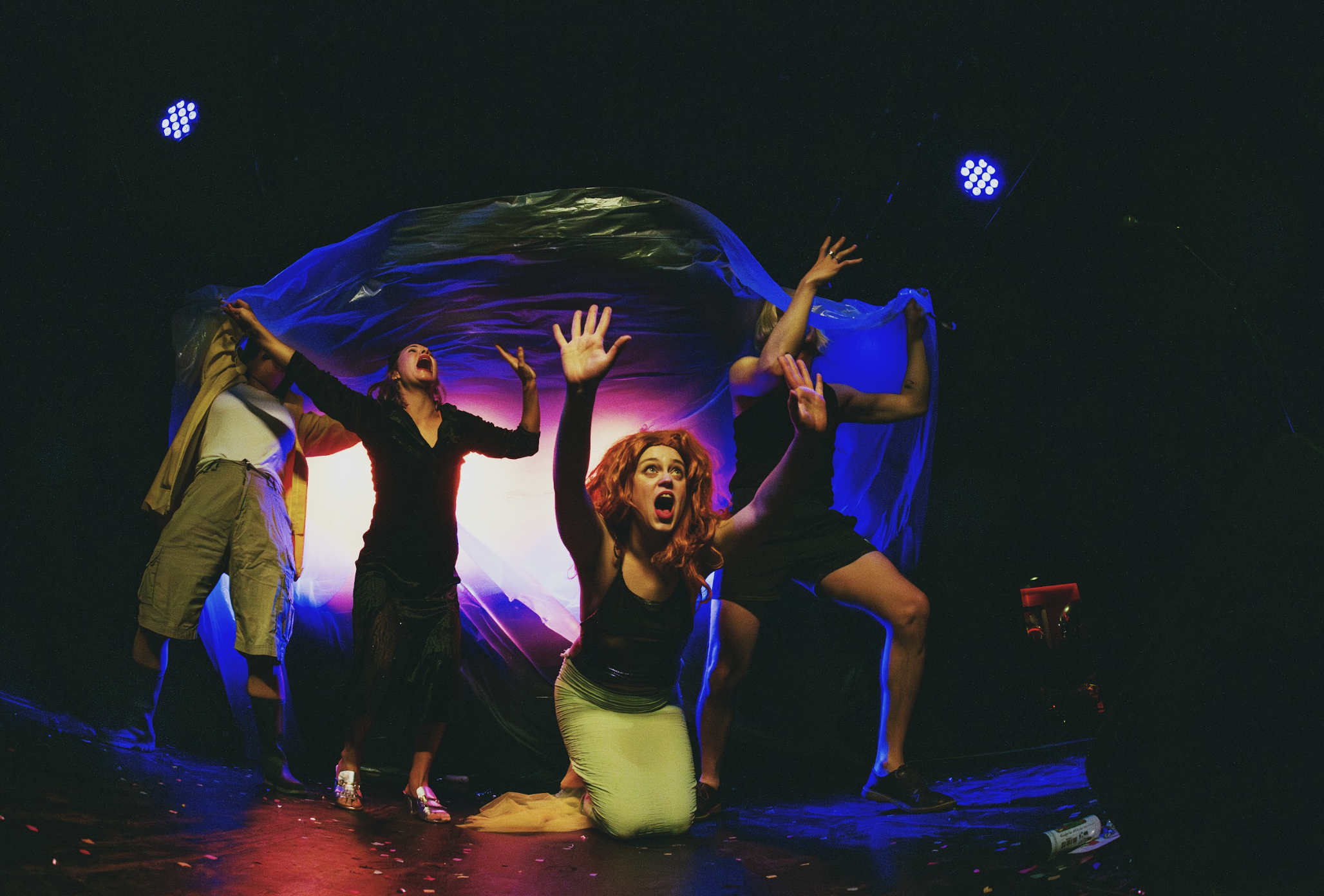 Four women on stage with expressive gestures, kneeling and holding a fabric backdrop, under colorful stage lighting, in a theatrical performance.