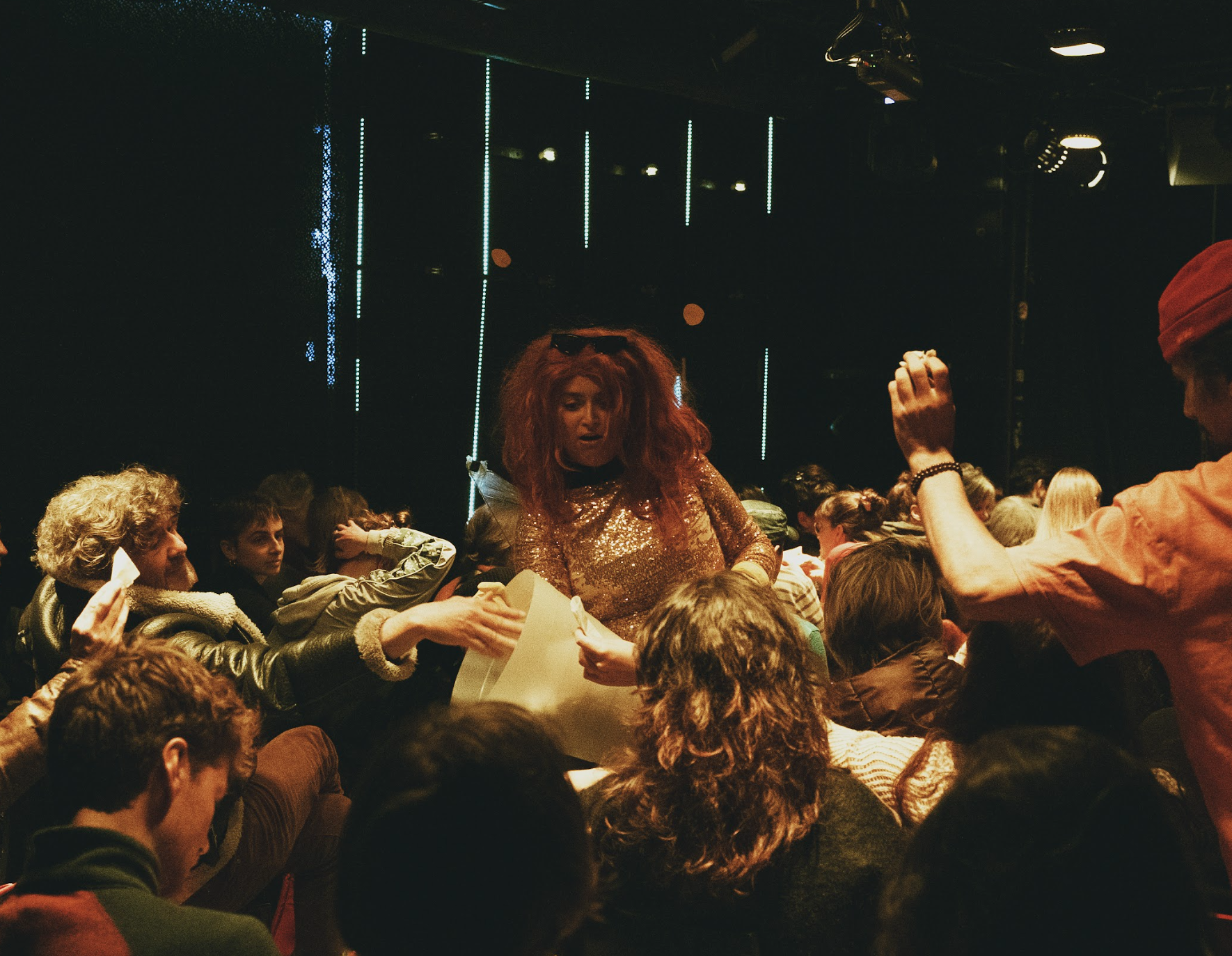 A crowded indoor event with a performer dressed in a shiny gold top and red wig, handing out paper to attendees. The background features vertical blue and green light strips.