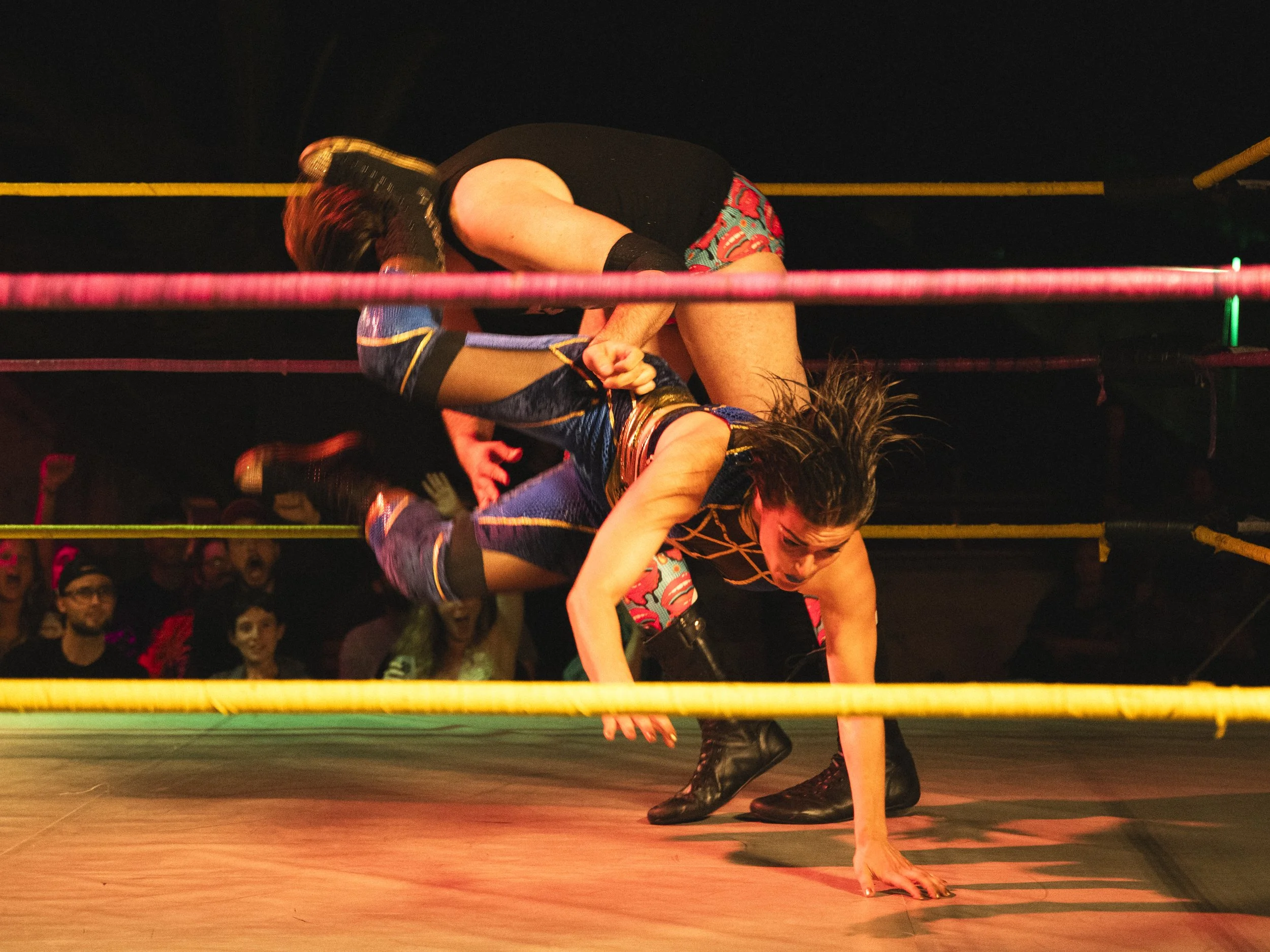 A female wrestler in colorful attire is on all fours on the wrestling ring, with her opponent grabbing her waist from behind during a match in front of an audience.