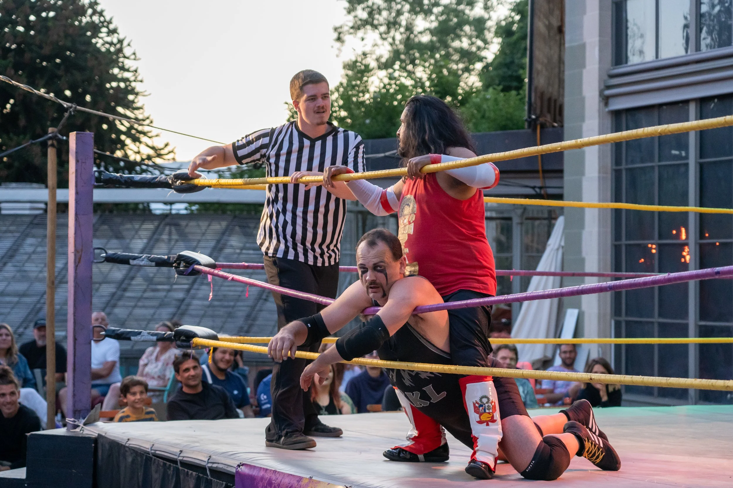 Two wrestlers in a ring, one on top of the other, while a referee observes. The wrestler on the bottom is on their hands and knees, and the wrestler on top is sitting. The referee is standing nearby, and there are spectators watching.