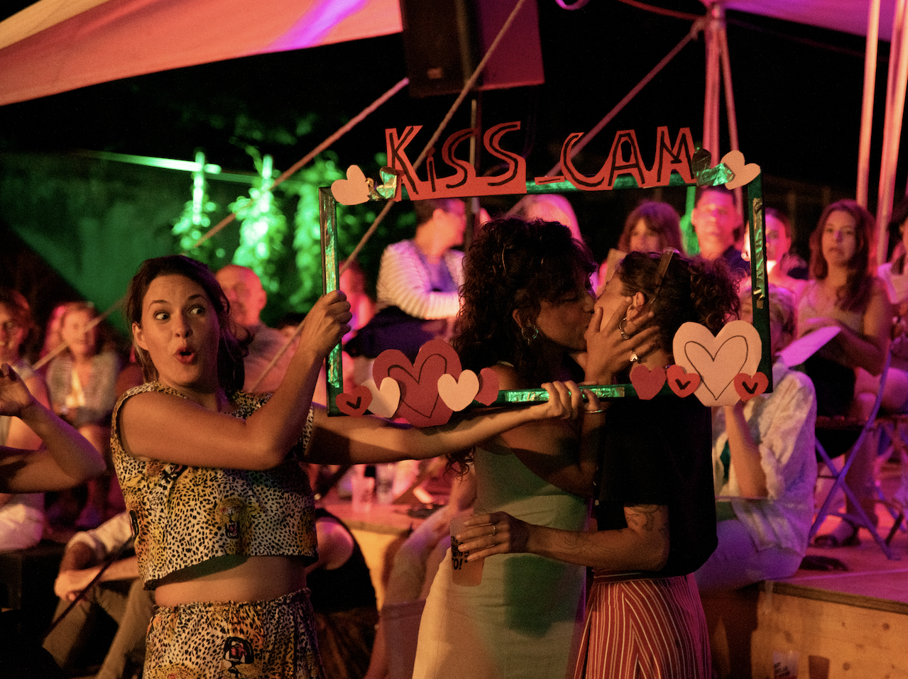 Two women sharing a kiss through a decorated photo frame that says 'KISS CAM,' held up by a woman with a surprised expression in the foreground. The scene is lit with colorful party lights, and people are sitting in the background.