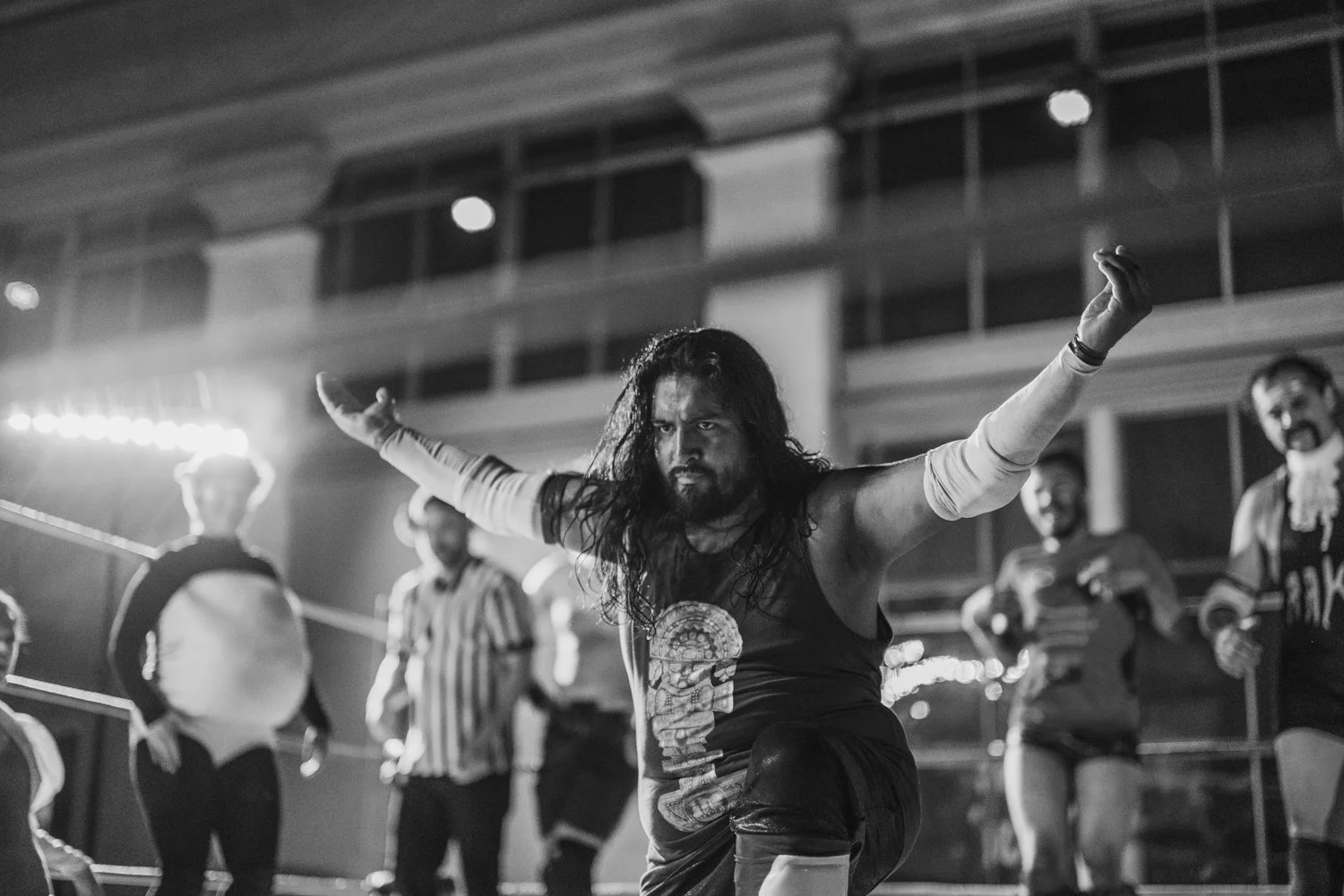 A black and white photo of a man with long hair and a beard, striking a dramatic pose with arms outstretched. The scene appears to be a dance or performance on stage with several other people in the background.