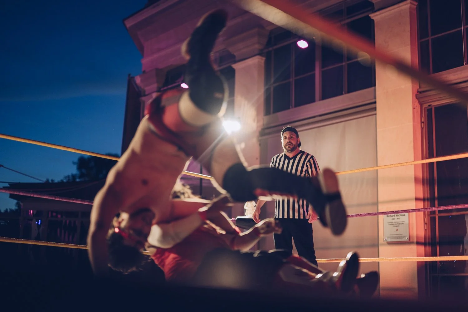 A wrestling match taking place outdoors at night with a wrestler on the ground and another wrestler in red and white attire in mid-air. A referee in black and white striped shirt observes the match, with a building in the background.