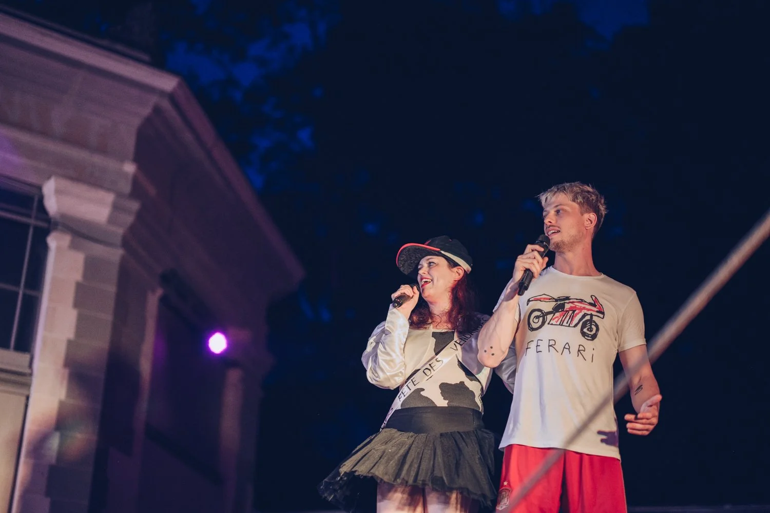 Two young people stand on a stage at night, holding microphones and speaking or singing. The woman has long dark hair, a baseball cap, a beige jacket, and a black skirt. The man has short blonde hair, a beige T-shirt with a motorcycle drawing and the