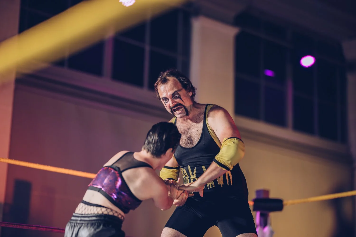 A wrestling match with a man wearing a black and yellow wrestling singlet forcing his opponent against the corner of the ring, both inside a wrestling ring with yellow ropes, in a gym.