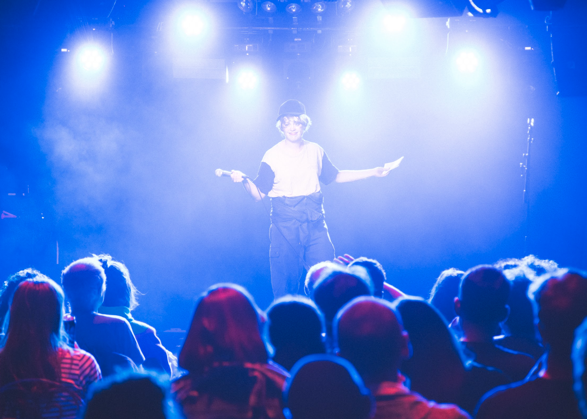 Performer on stage holding microphone and paper, wearing hat, with audience watching, stage illuminated with bright blue and white lights.