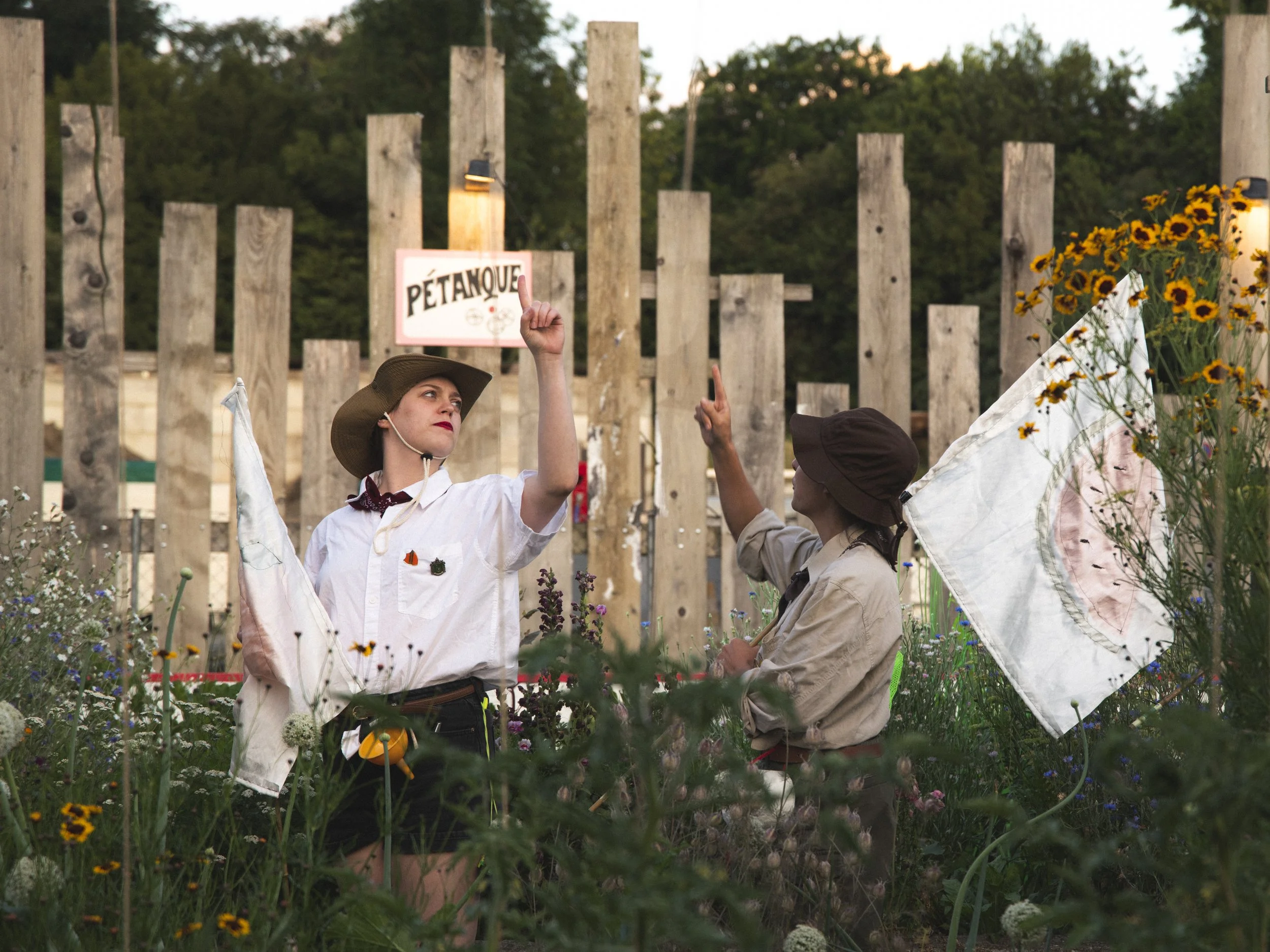 Two women dressed in vintage clothing and hats, engaging in a conversation amid tall flowers. One woman holds and gestures with a finger pointing upwards, while the other appears to be listening. There is a wooden fence and a sign in the background.
