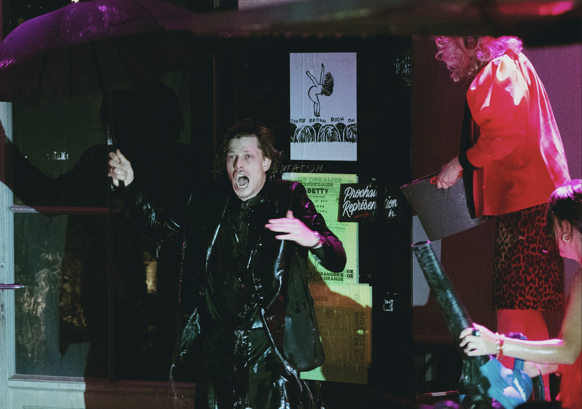 A man with curly hair wearing a shiny black suit is gesturing wildly with his mouth open, inside a glass display case. Beside him, an older person in a bright pink jacket with curly hair, holding a silver bag, is partially visible. The background has