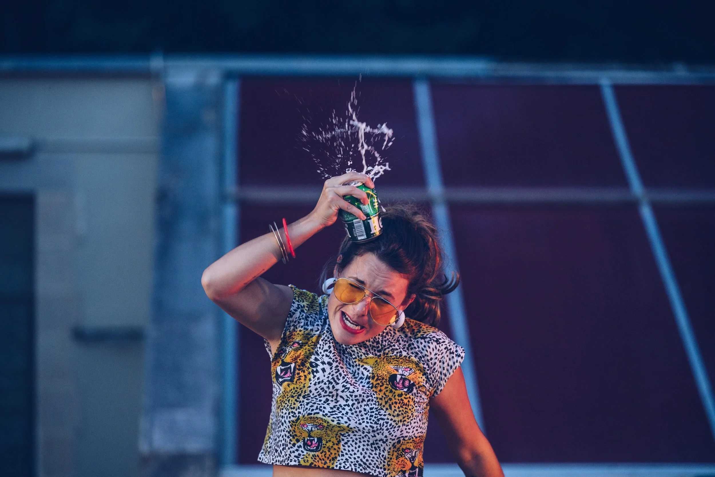 A woman with dark hair, wearing yellow sunglasses and a tiger print top, is holding a can of soda on her head as it spills. She has an expression of surprise or discomfort.