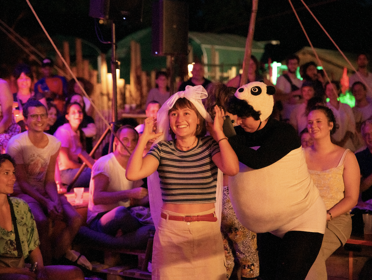 A woman and a person in a panda costume dance and enjoy a lively, colorful event or party at night with a crowd in the background.