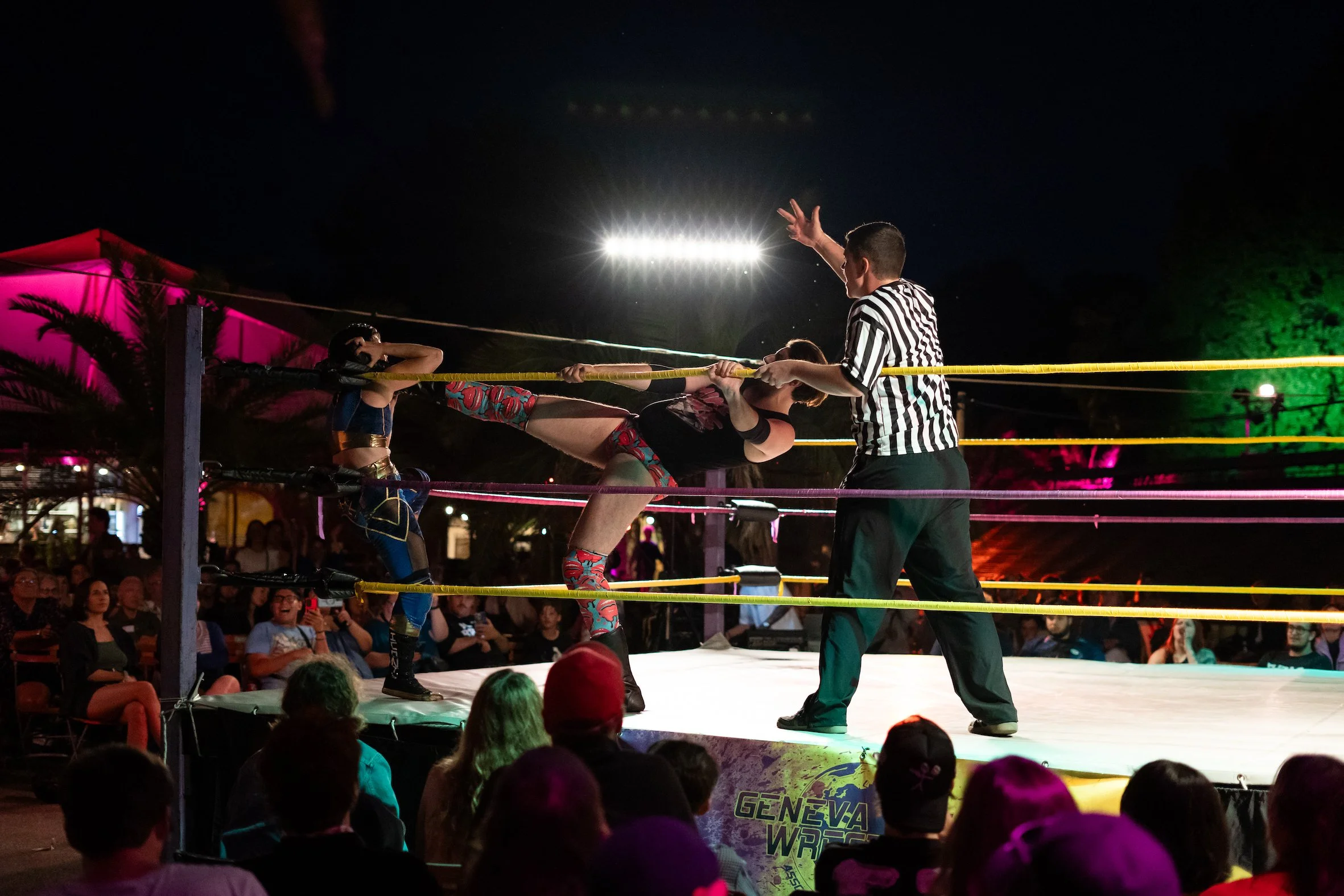 A wrestling match taking place outdoors at night, with a female wrestler delivering a high kick to her opponent, who is blocking. The referee is observing closely, and an audience watches from around the ring, illuminated by colorful lights.