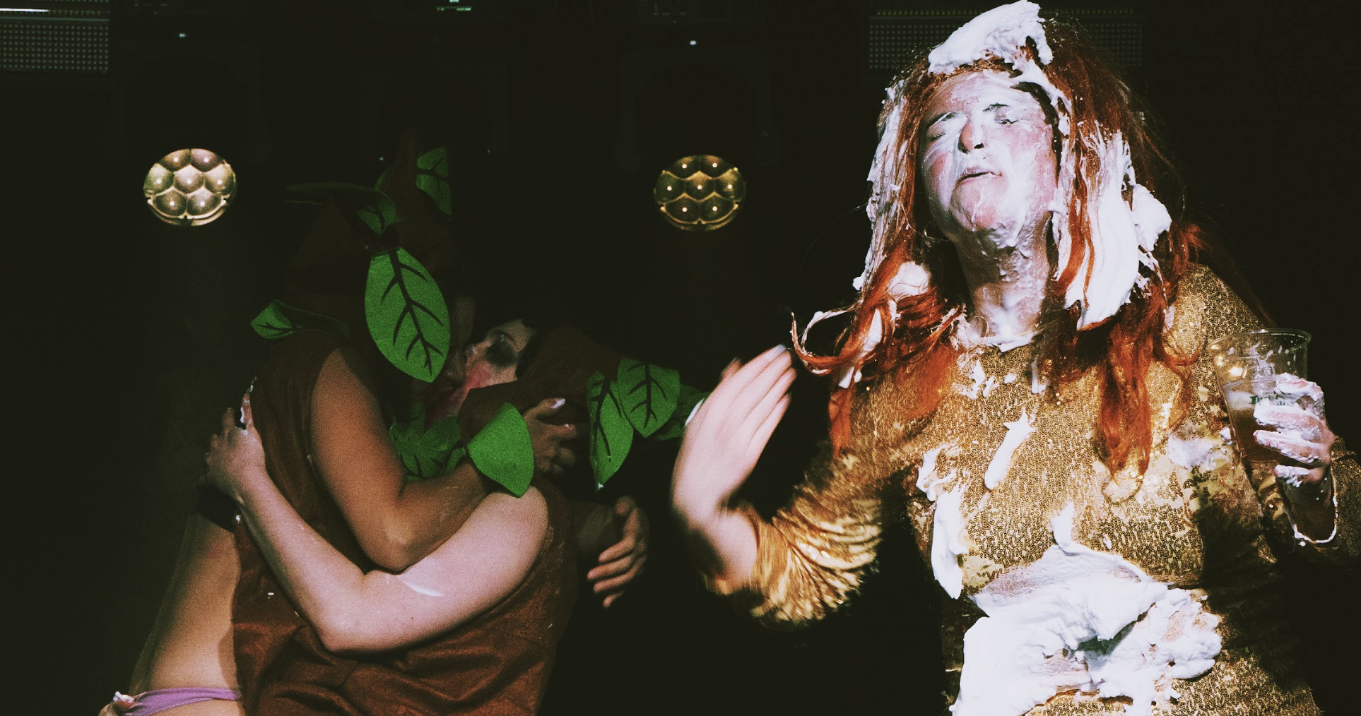 People dressed in costumes at a party, with one person covered in cake and whipped cream, wearing a gold dress, and others in leaf costumes.