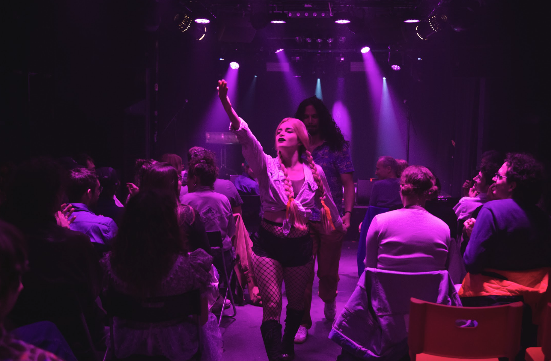A female performer with long braided hair and fishnet stockings on stage under purple lights, raising her right arm, while a diverse audience watches her at a concert or show.