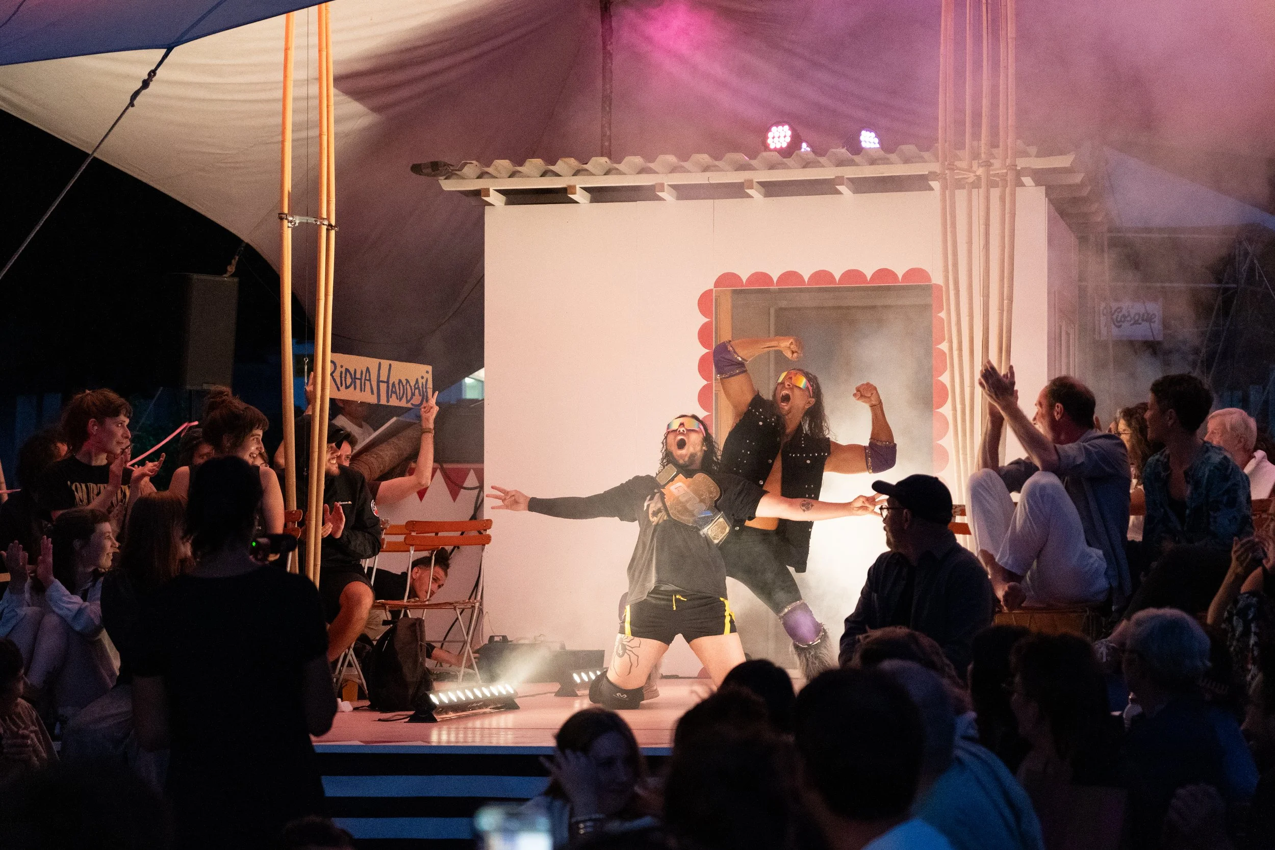 Performers with sunglasses posing dramatically on stage at a circus show, surrounded by audience members and colorful stage lights.