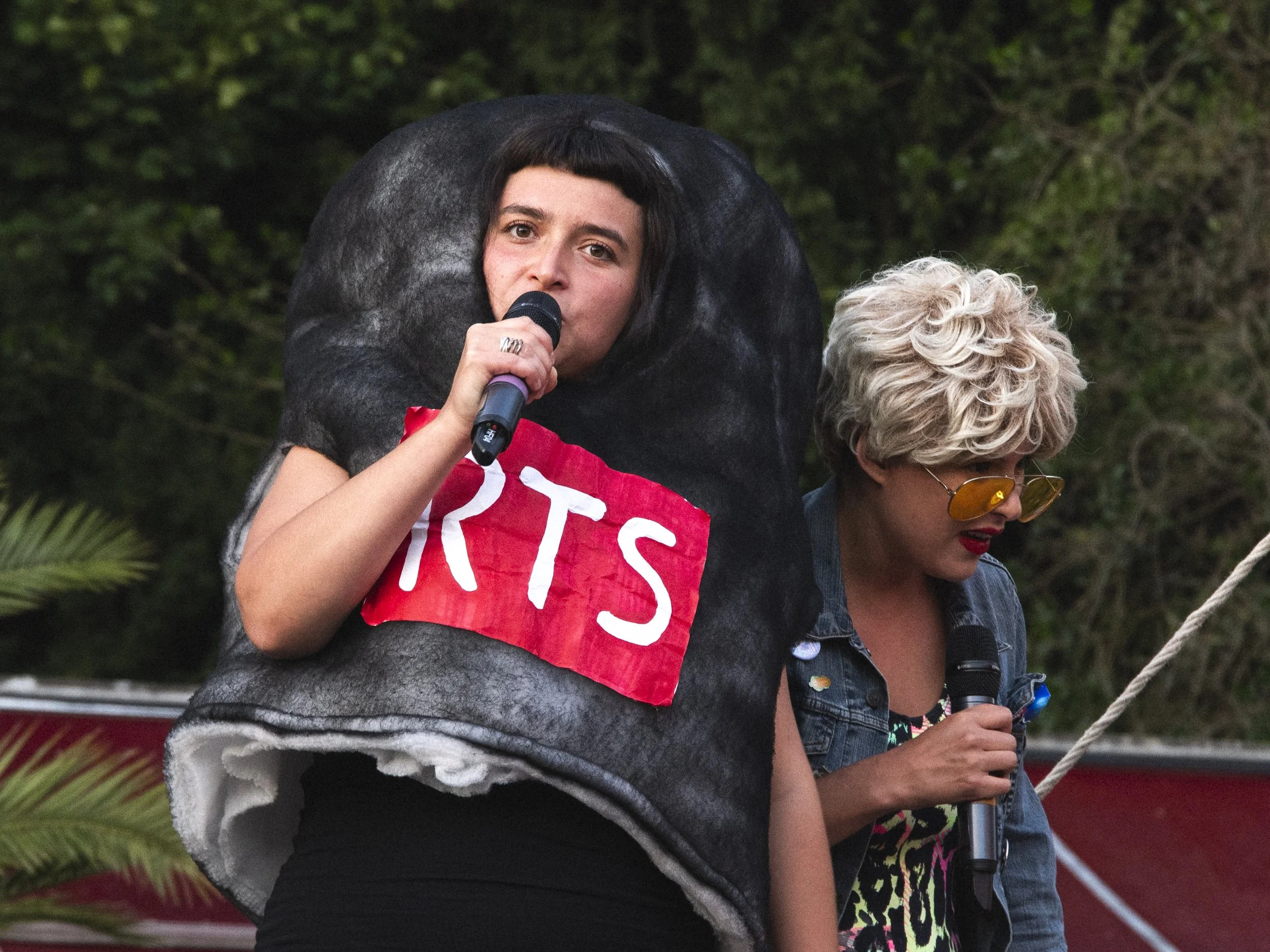 Two women are on stage, speaking into microphones, with one dressed in a costume resembling a giant black and gray animal with a red sign reading 'ARTS' on it. The woman in the costume has short dark hair with bangs, and the other woman has curly blo