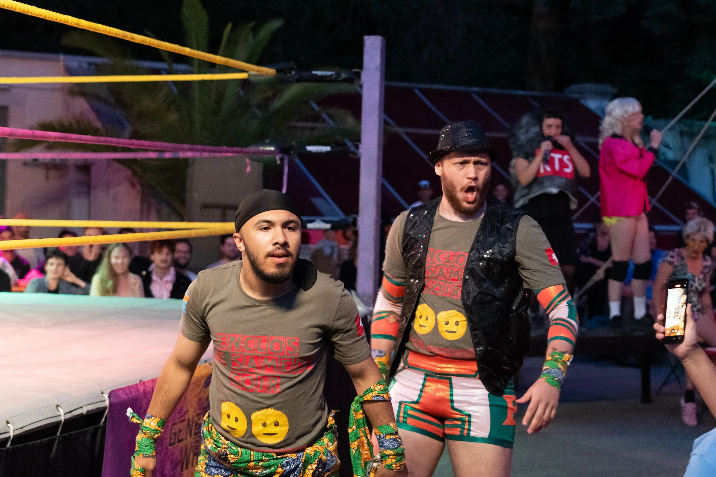 Two men in colorful wrestling attire stand inside a wrestling ring during an event, with an audience in the background.