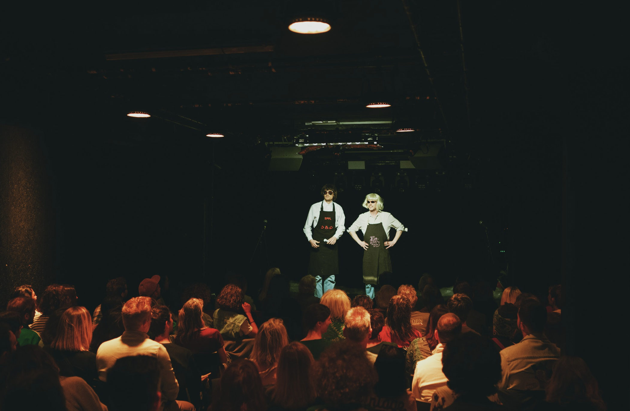 Two performers on stage dressed as chefs, standing with their hands on their hips, wearing aprons and sunglasses, in front of an audience in a dark theater.