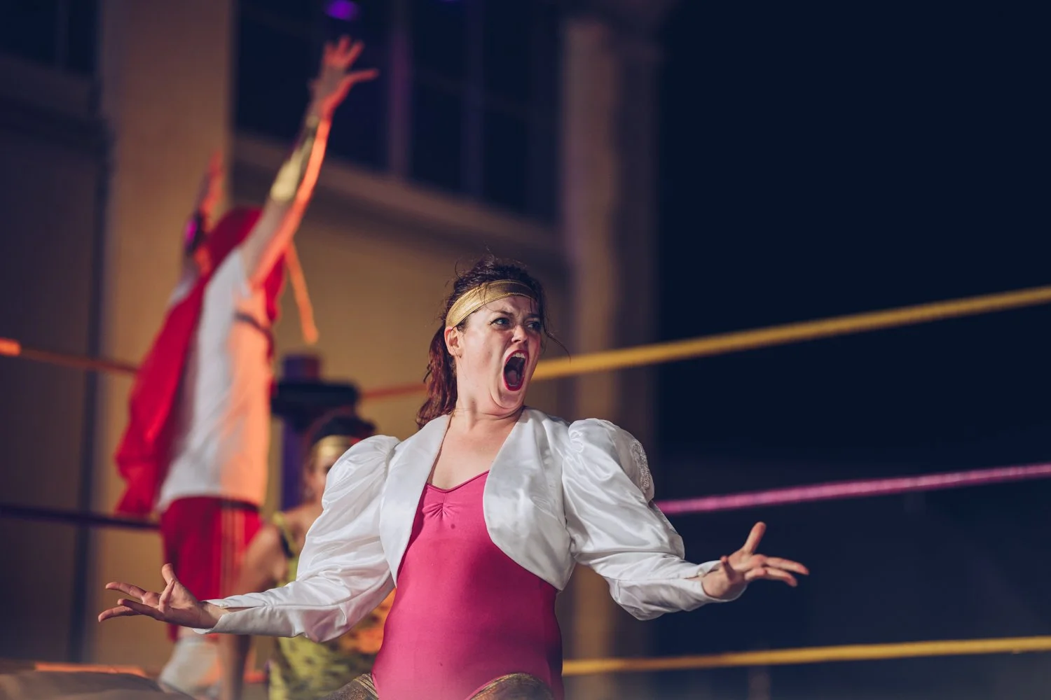 A woman with a headband and pink top, in a wrestling ring, appears to be yelling or shouting, with an arm raised in the background.