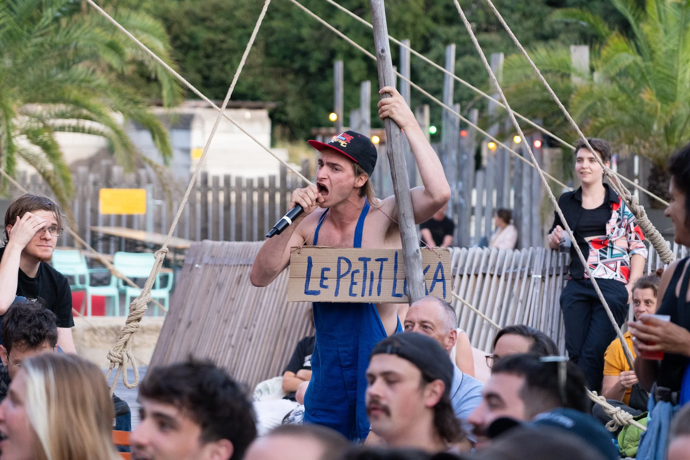 A man with long hair and a cap holding a microphone and a sign that reads 'LE PETIT LALA' stands on a boat or dock, speaking passionately to a group of people gathered around him. The background features palm trees and a wooden fence.