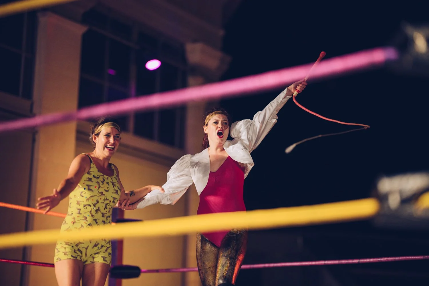 Two women celebrating in a wrestling ring, one holding a pink whip and the other smiling, at night outdoors.