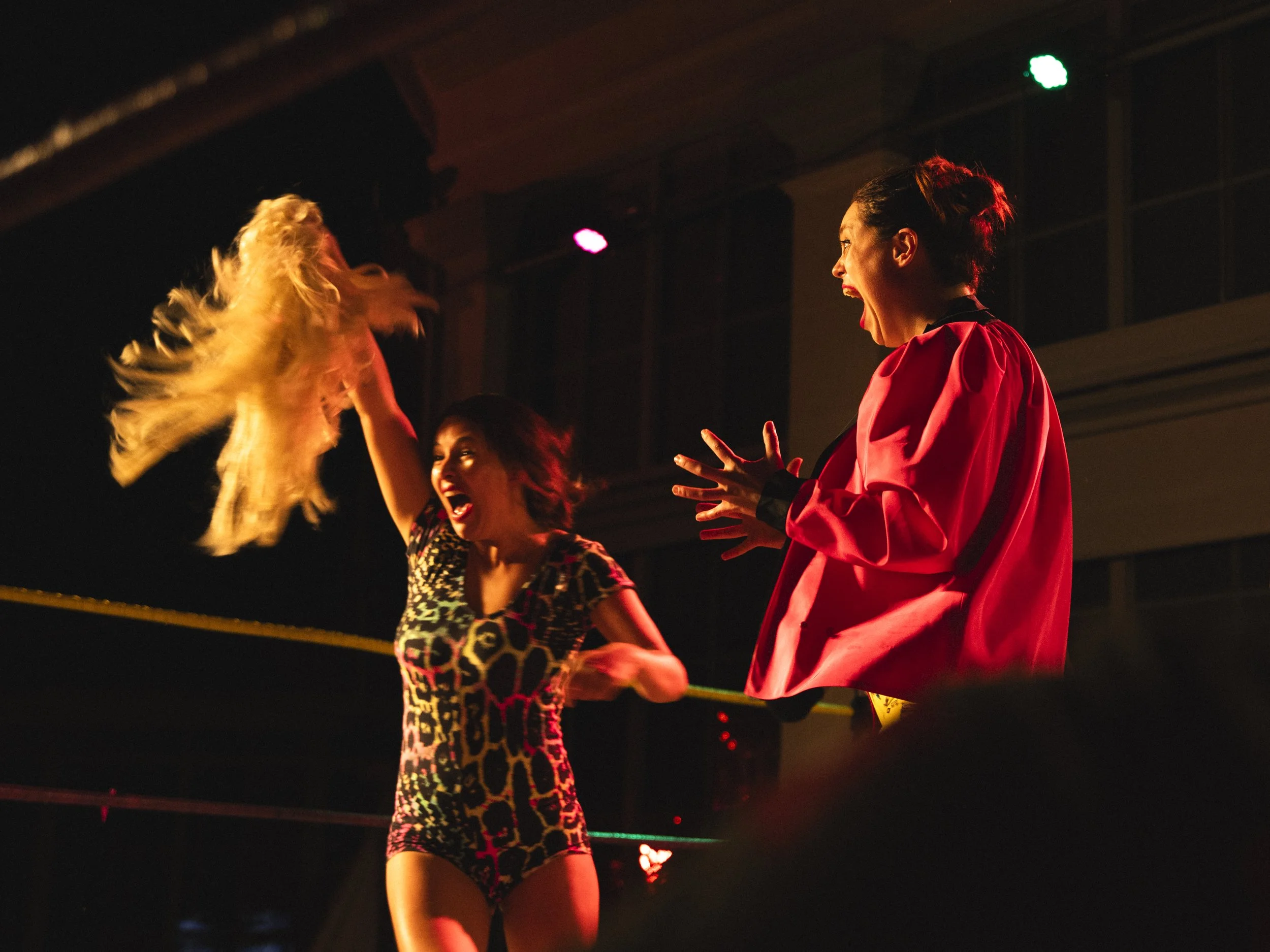 Two women excitedly celebrating in a wrestling ring during a match. One woman is wearing a multicolored animal print outfit, waving her blond hair, and the other woman is wearing a red shirt, expressing shock or excitement.