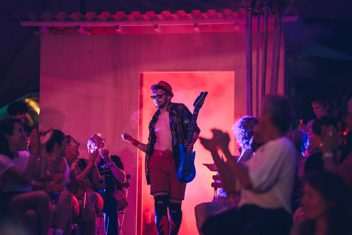 A performer dressed in colorful, eclectic clothing with a guitar, hat, and sunglasses, walking through a lively audience in a dimly lit, vibrant setting with pink and purple lighting.