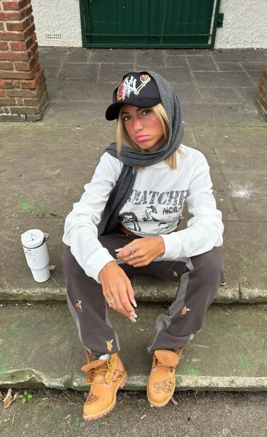 A young woman sitting on a concrete step outdoors, wearing a baseball cap, a scarf around her neck, a white sweatshirt, distressed gray pants, and tan boots, with a water bottle on the ground beside her.