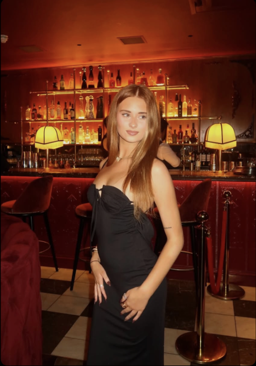A young woman with long light brown hair and a black strapless dress stands in a dimly lit bar with a red background and bottles of alcohol on shelves behind her.