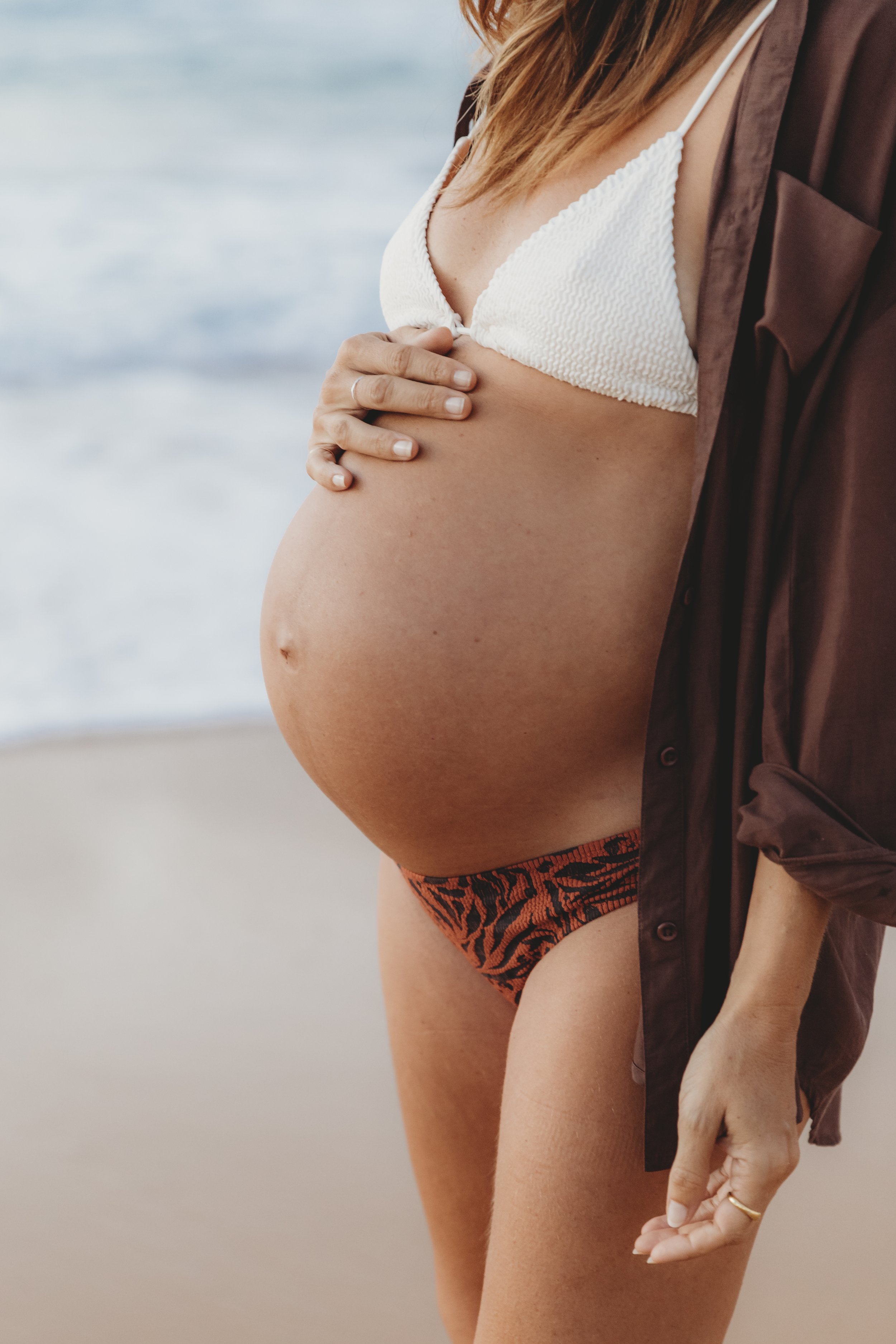 Pregnant woman standing on the beach, wearing a white bikini top, orange and black animal print bikini bottoms, and a brown shirt draped over her shoulders, with her hand resting on her belly.