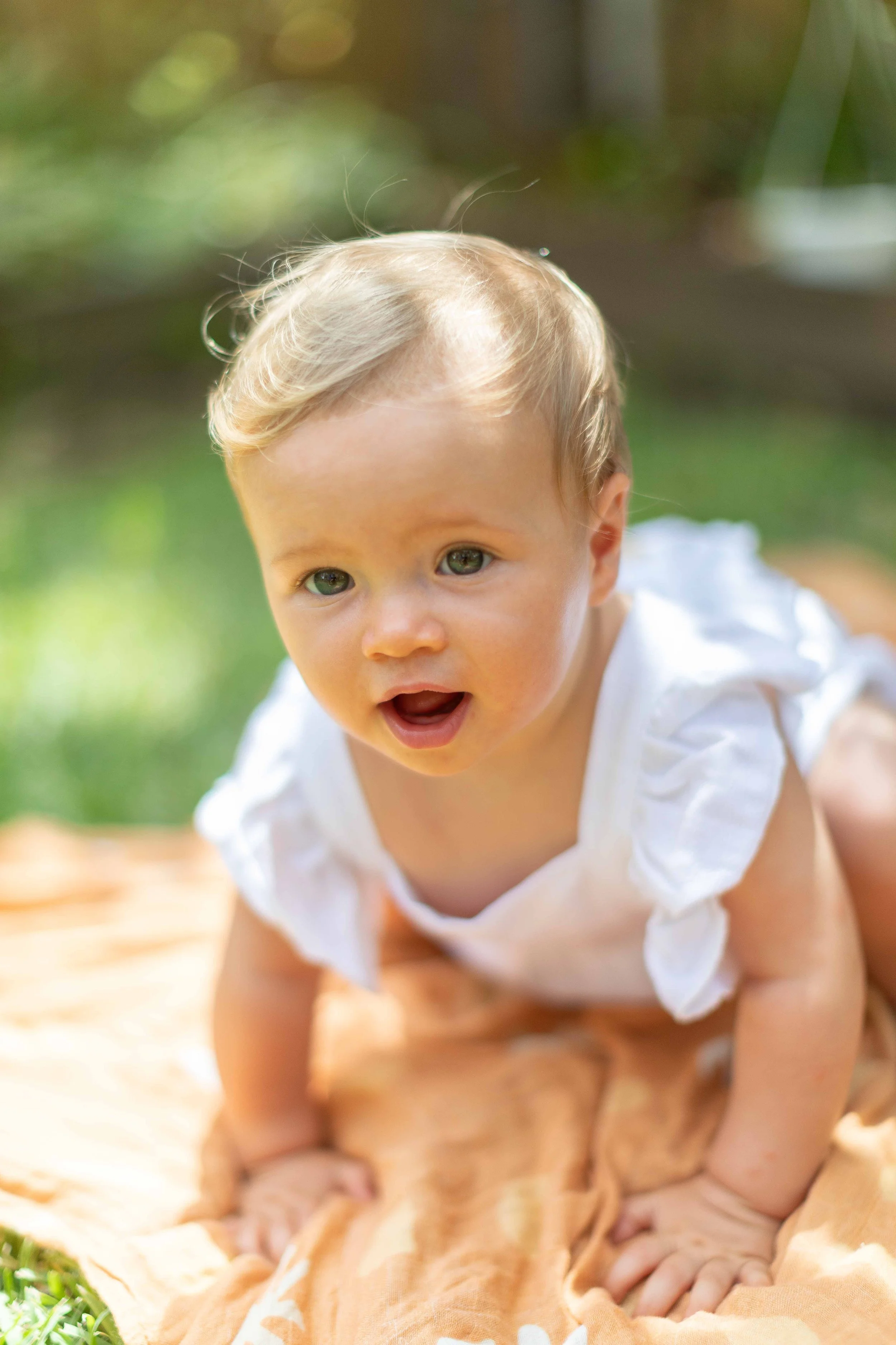 A toddler crawling on a blanket outdoors, looking at the camera with an open mouth and expression of curiosity.