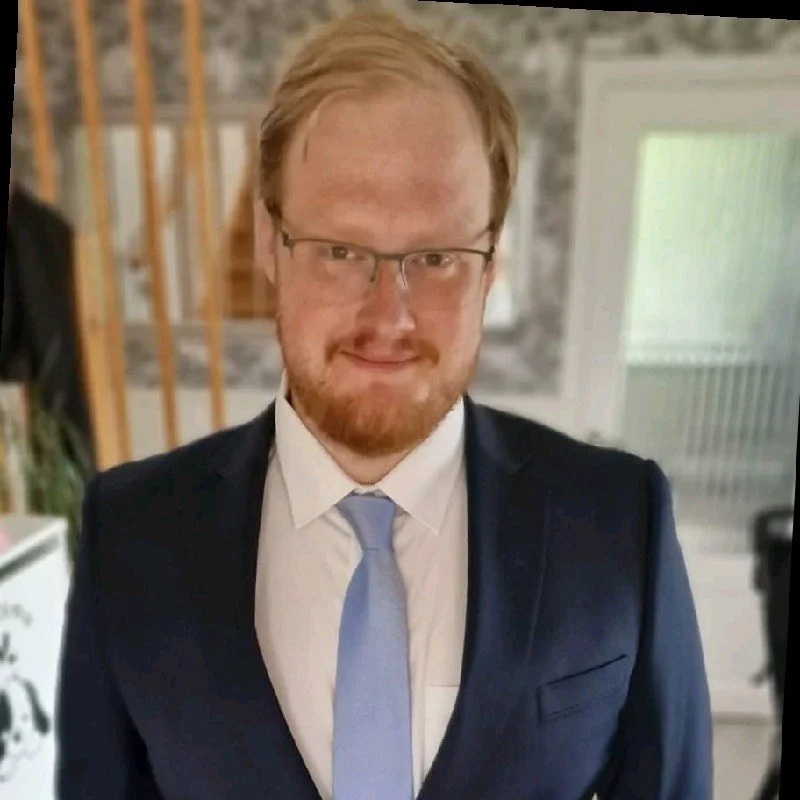 A young man with a beard and glasses standing outdoors, looking at the camera, with a book store and cars in the background during sunset.