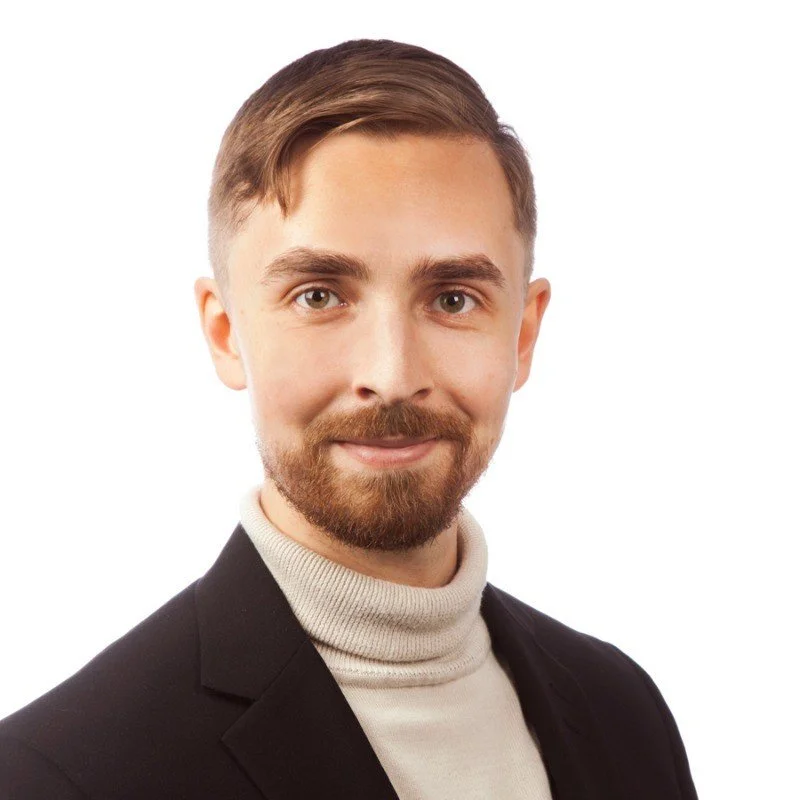 A man with curly dark hair, glasses, a nose ring, and a beard, wearing a dark sweater, looking slightly to the side with a gentle smile in an indoor setting.