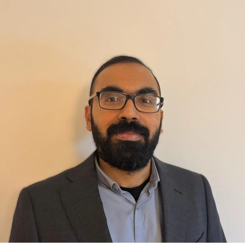 A woman with dark hair in a messy bun, smiling, with a nose piercing, wearing a white top, against a plain white background.