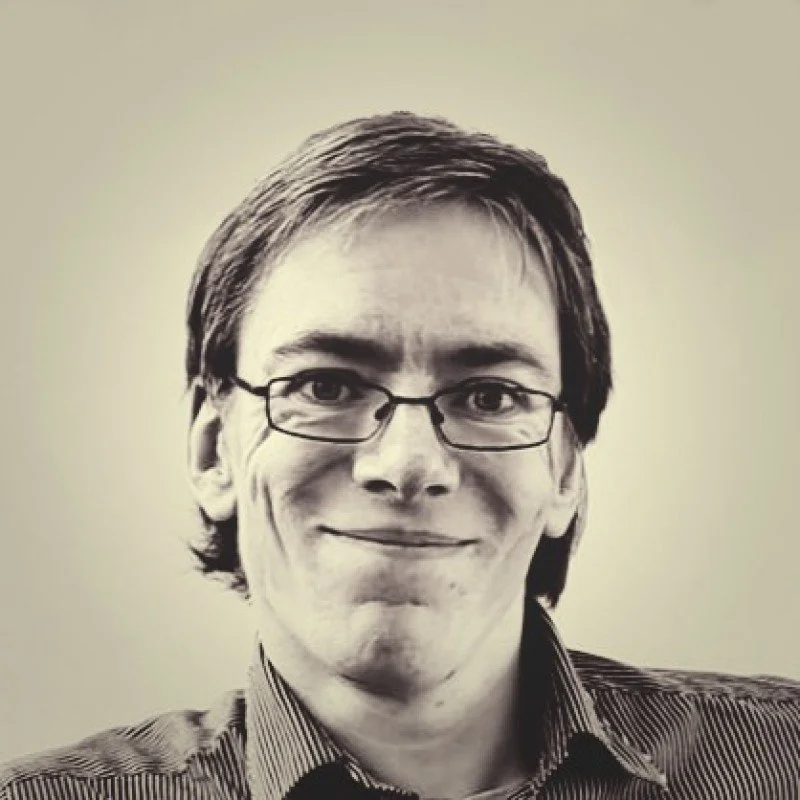 A young man with glasses and a beard standing outside near parked cars, with a building and a sign in the background.