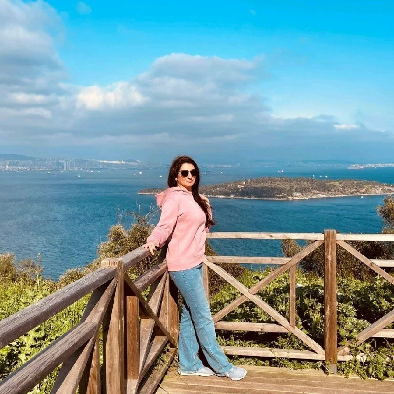 A woman with dark, messy hair styled in a high bun, smiling, wearing a nose ring and a white shirt, with a plain white background.