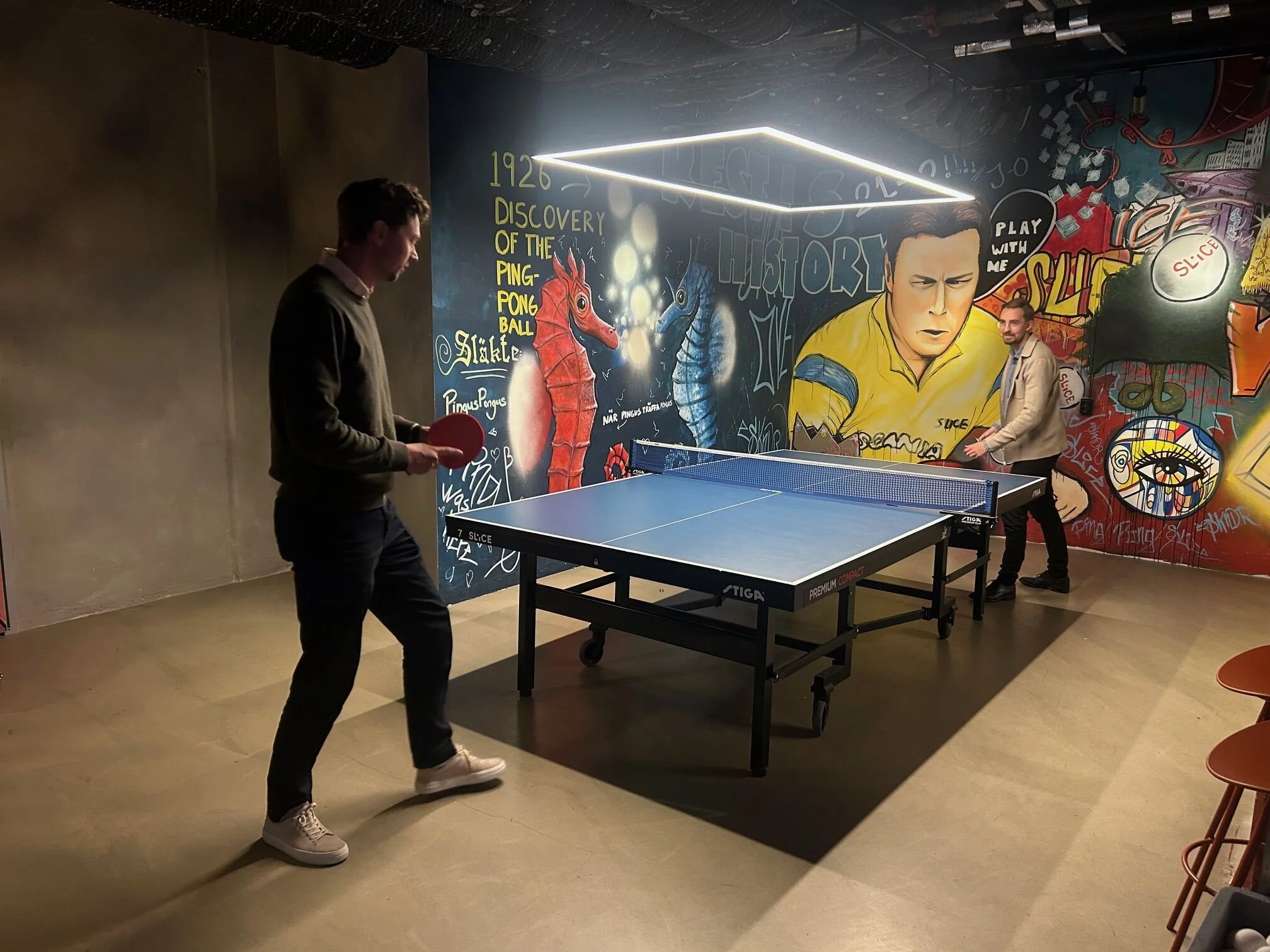 Two men playing table tennis in a room with colorful graffiti murals on the walls, including a cartoon character and text.